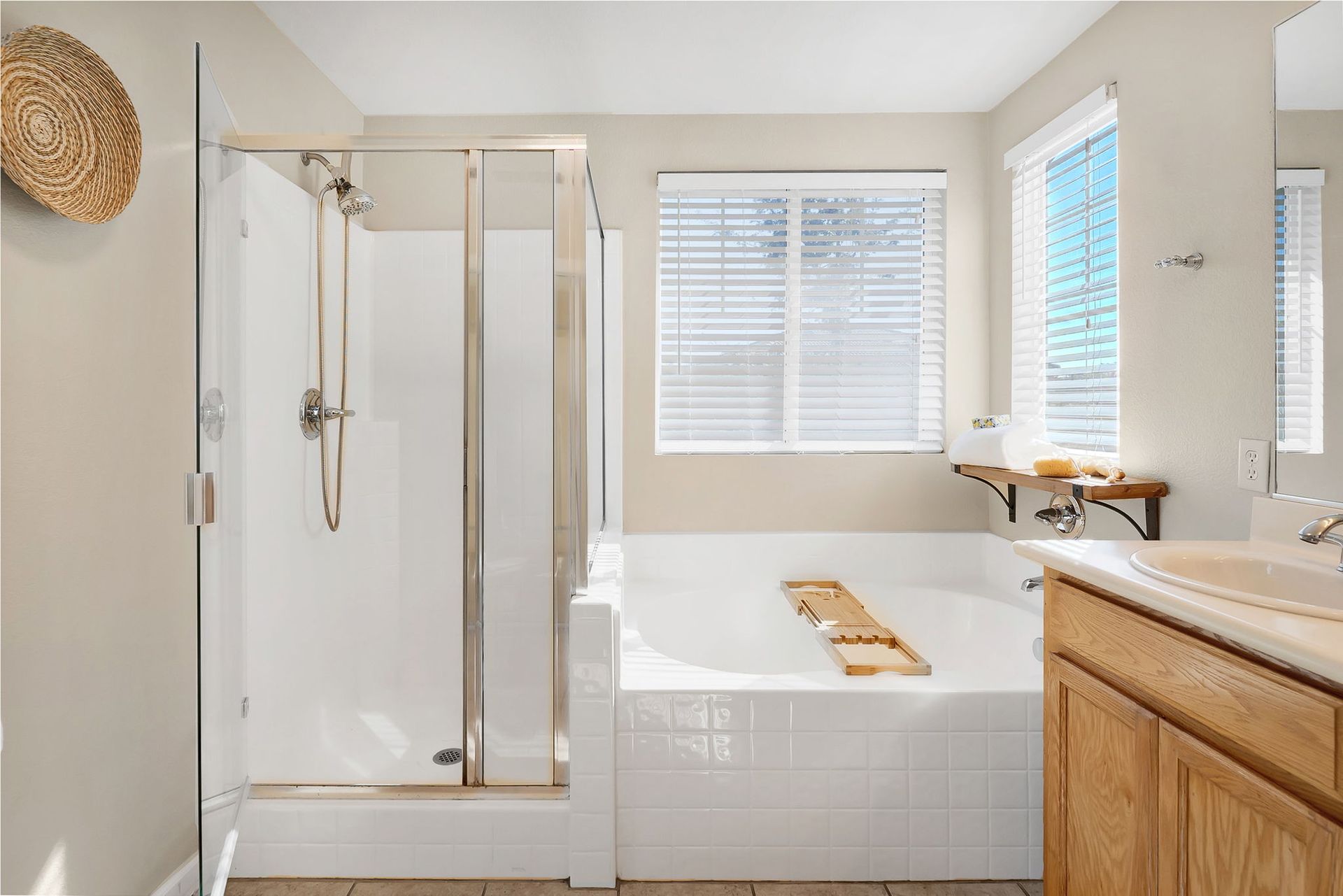 Bathroom with white shower and tub, wood vanity and woven wall decor. Sunlight streams through window.
