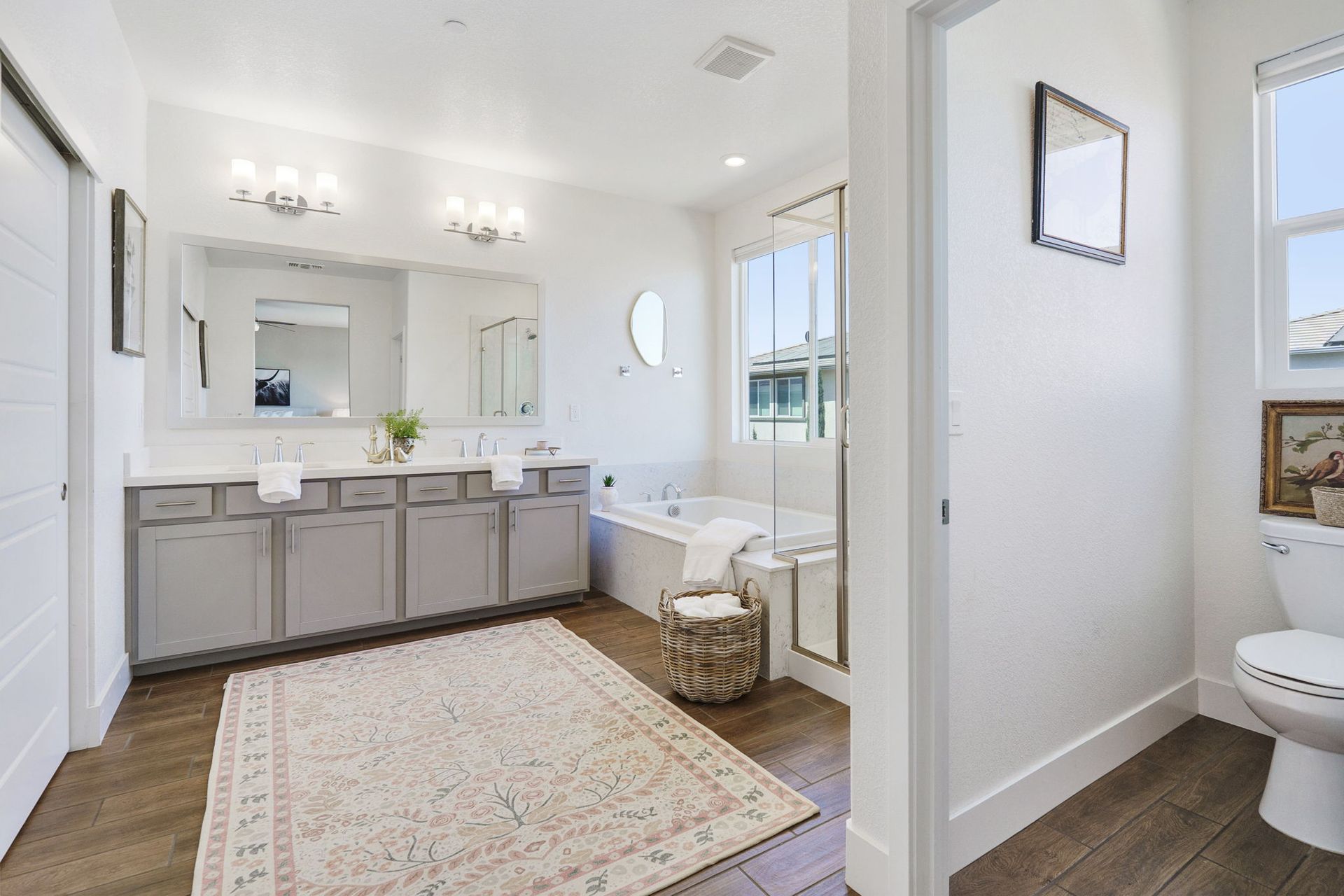 Spacious, bright bathroom with double vanity, bathtub, and toilet; light wood floor and area rug.