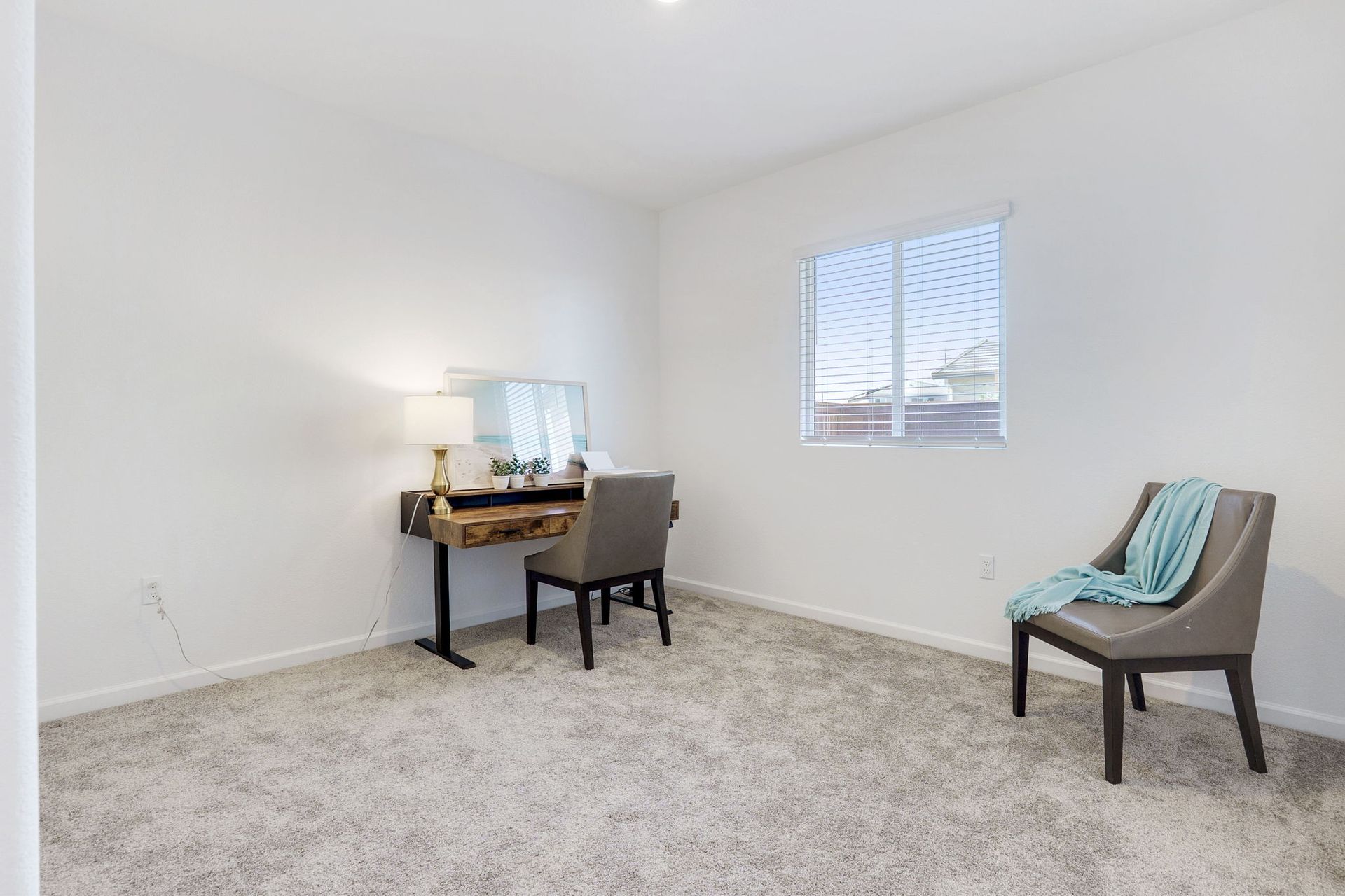A sparsely furnished room with a desk, chair, and armchair; soft carpet and a window with blinds.