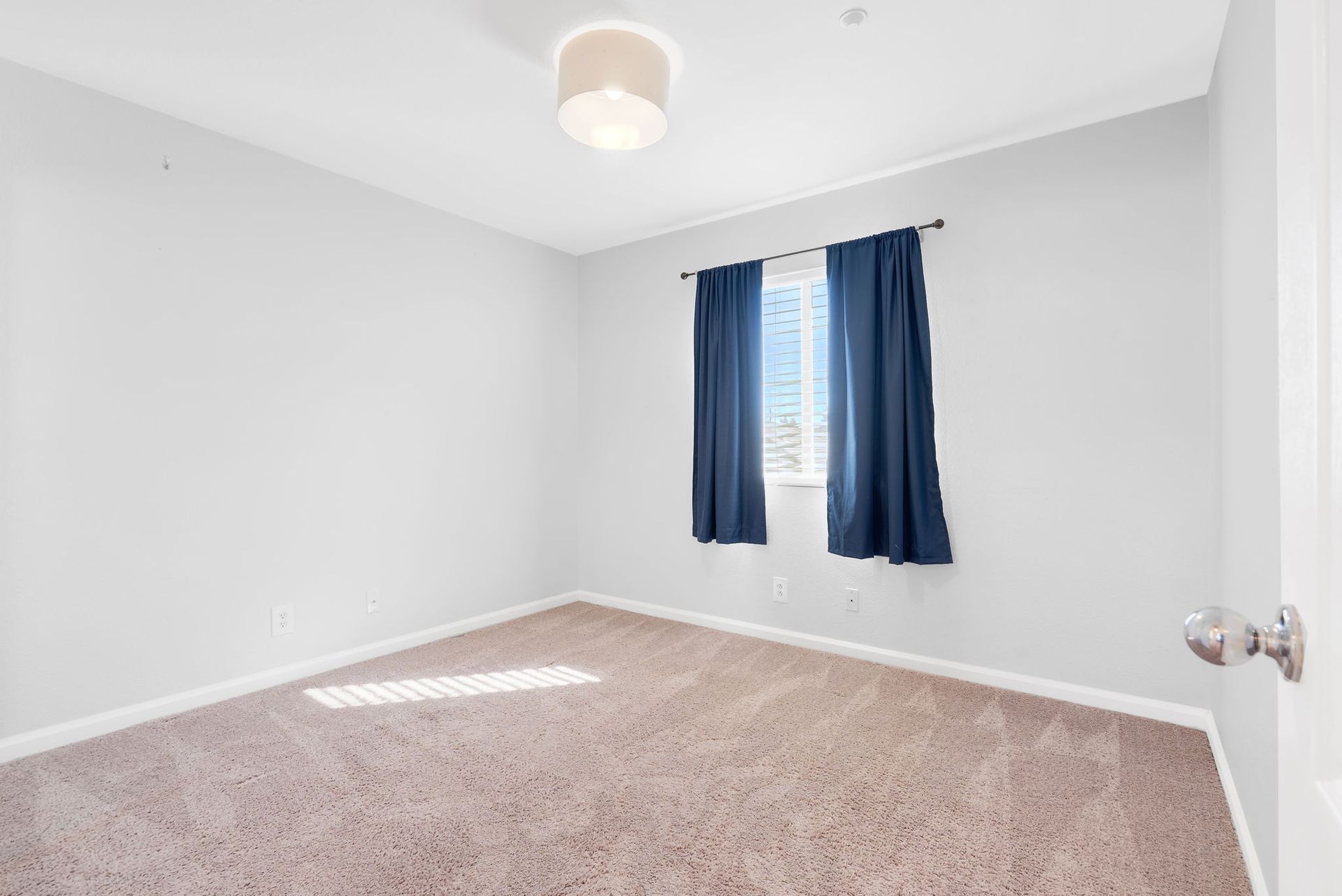 Empty bedroom with gray walls, beige carpet, navy blue curtains, and a white ceiling light.