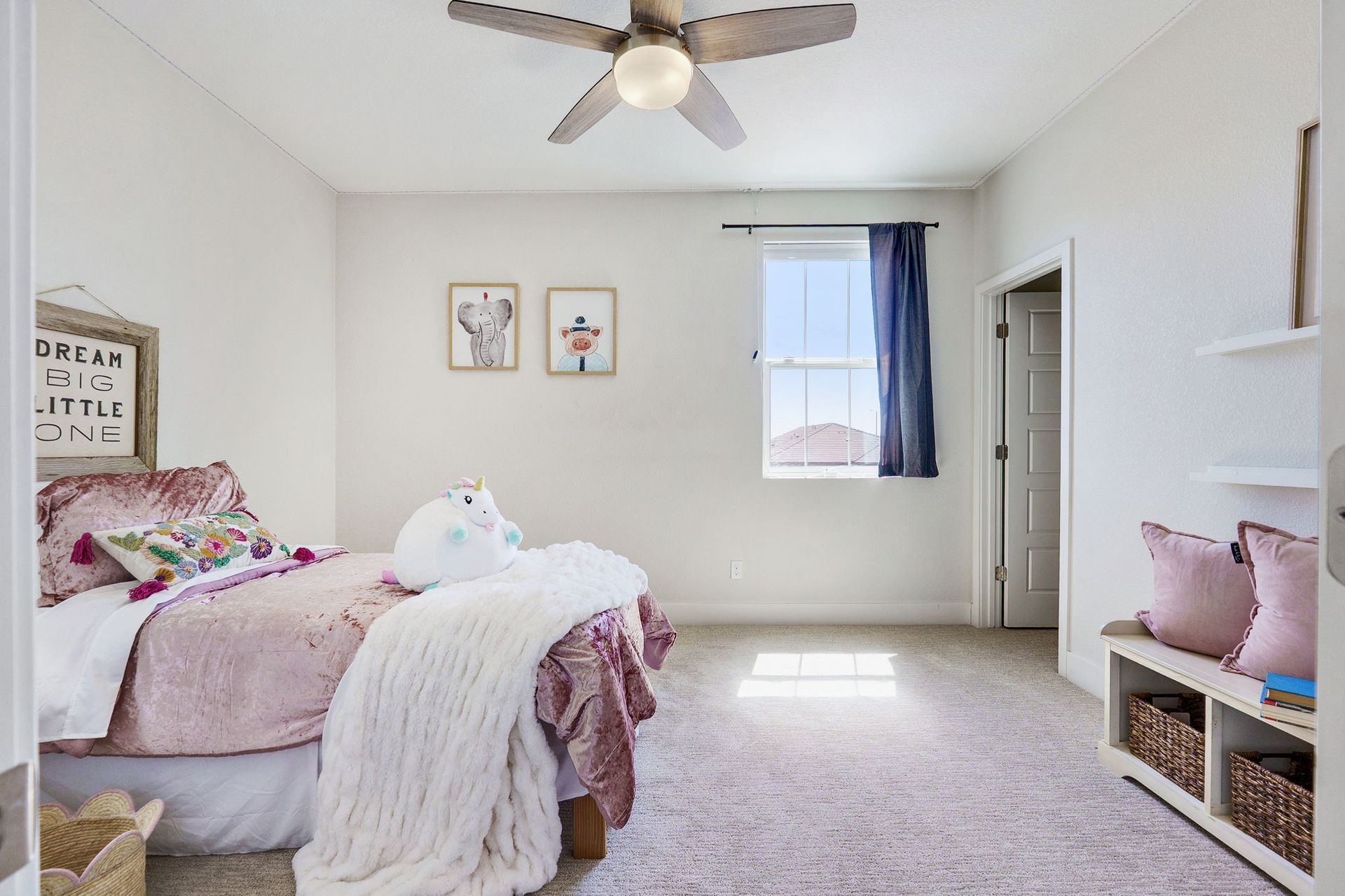 Girl's bedroom with pink bedding, two framed pictures, a window with blue curtains, and a white ceiling fan.