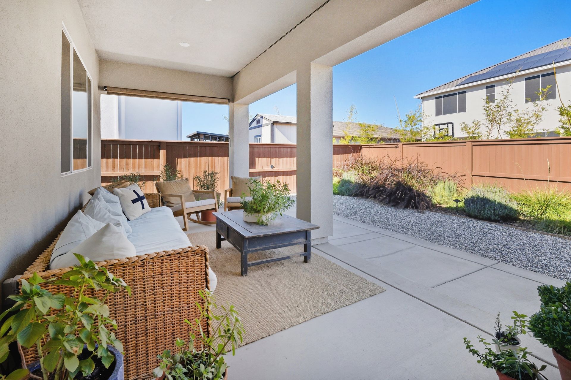 Outdoor patio with seating, coffee table, and greenery, overlooking a yard and houses on a sunny day.