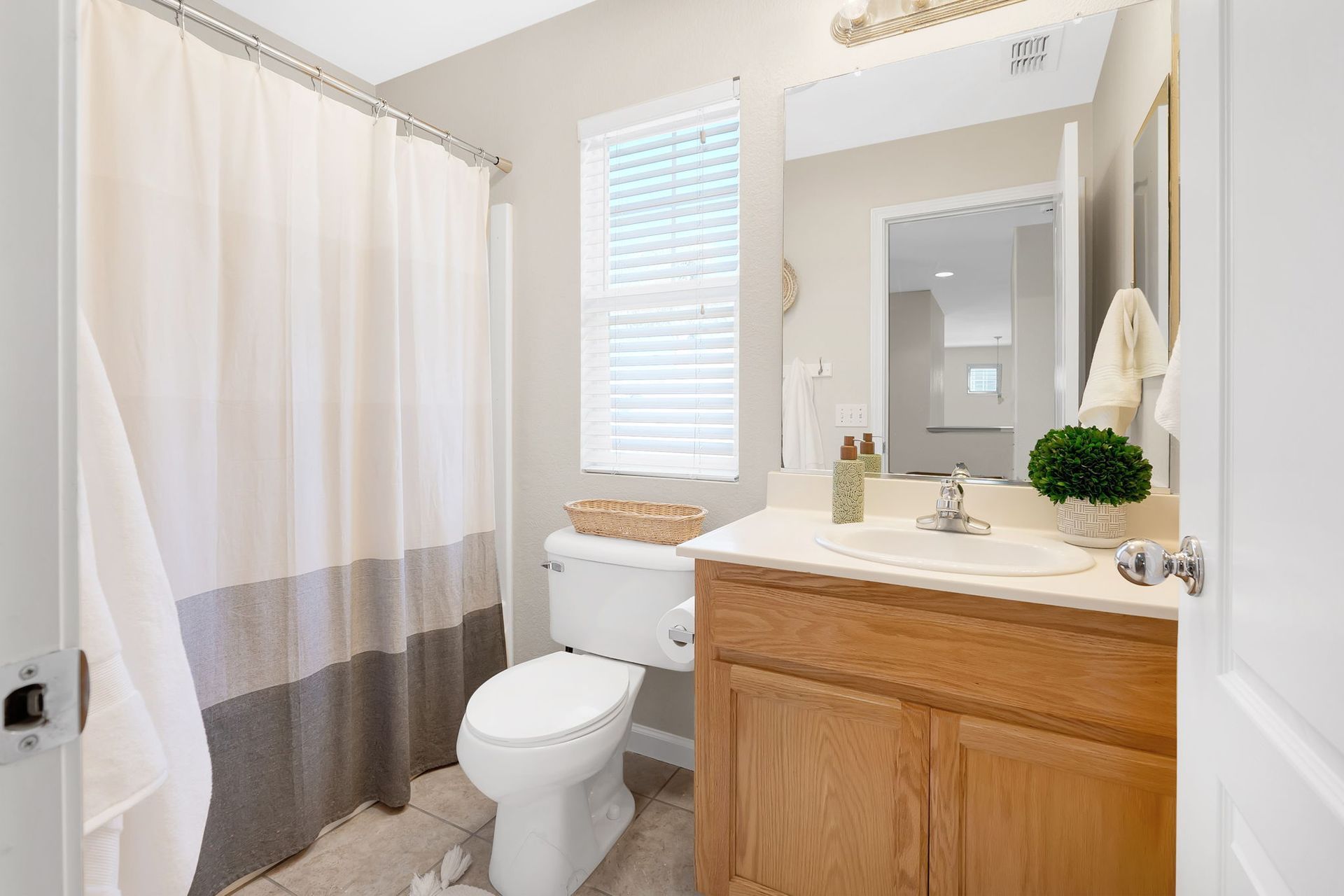 Bathroom with toilet, vanity, shower with curtain. Beige, gray, and white tones. Window with blinds.