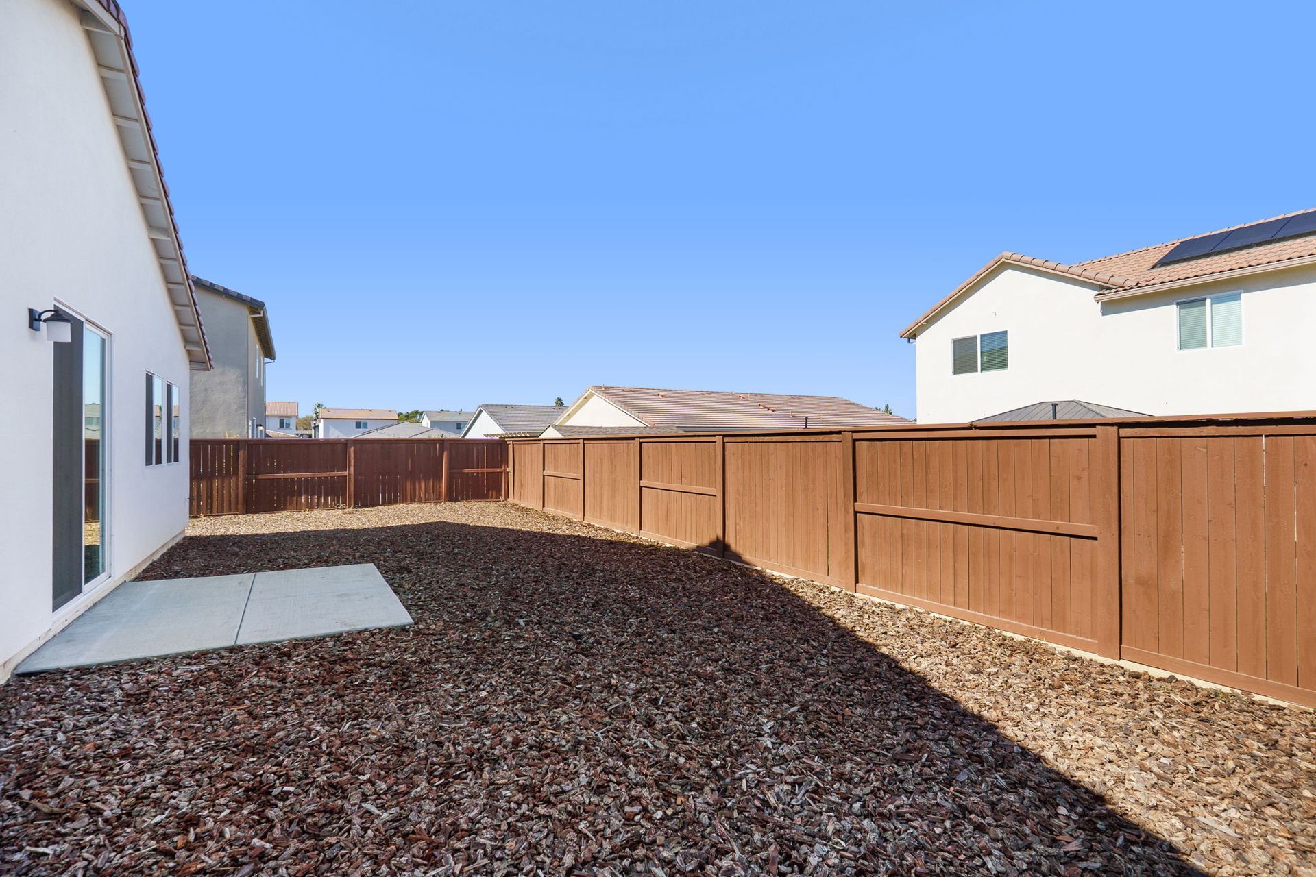 Backyard with brown mulch, concrete patio, and wooden fence. Houses and blue sky in background.