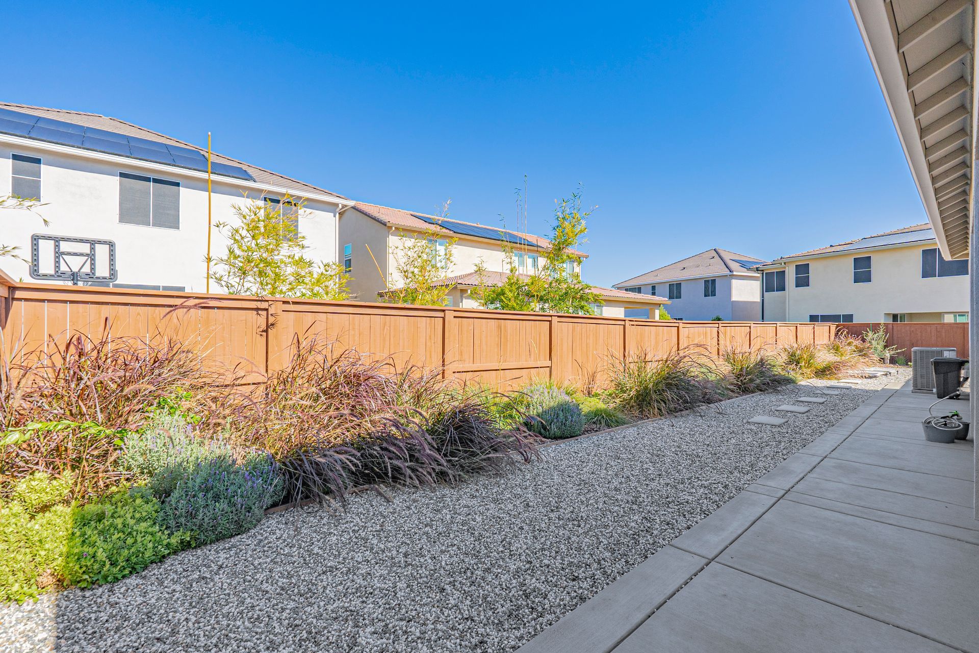 Backyard with a gravel path, wooden fence, and homes under a blue sky.
