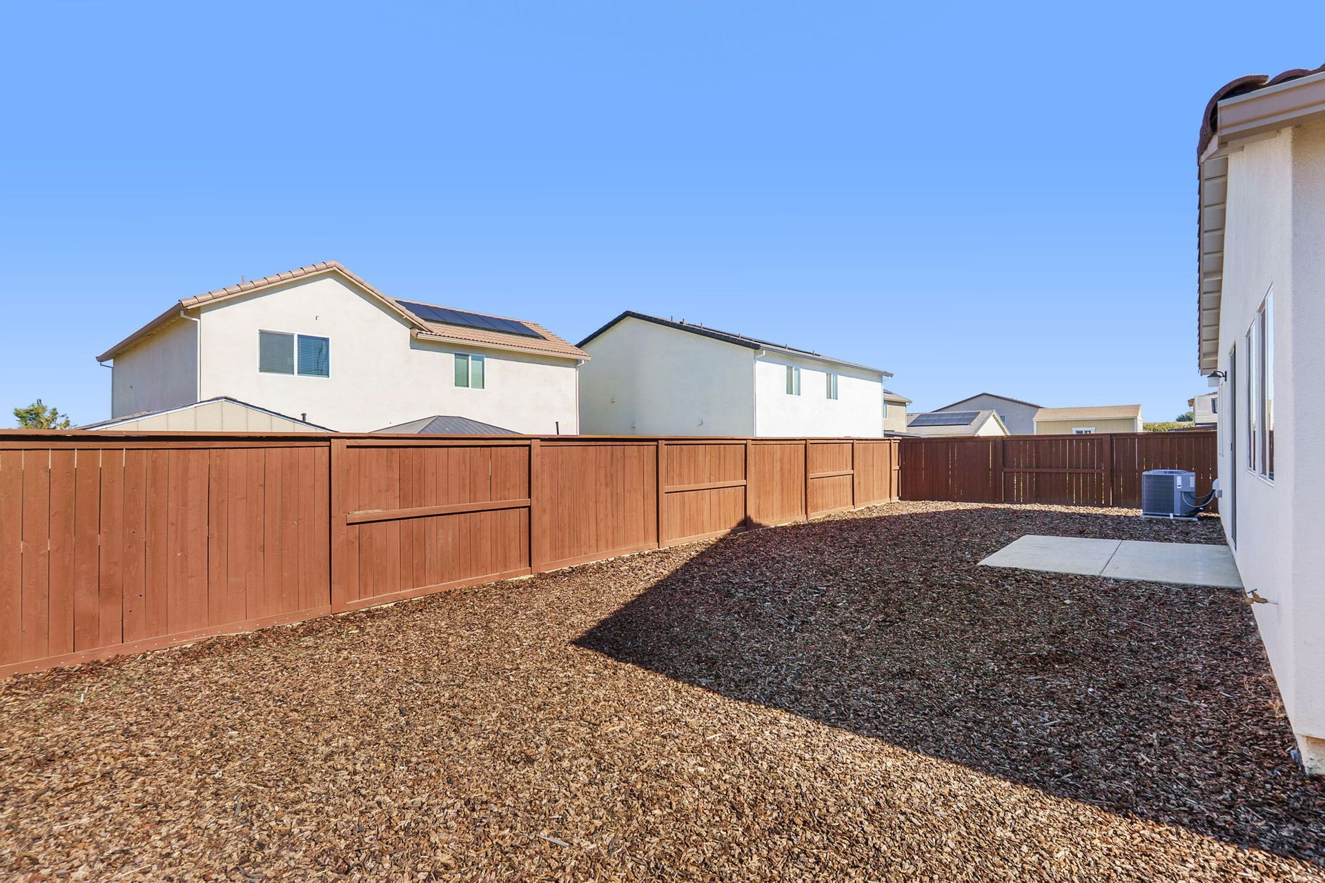 Backyard with brown gravel, wooden fence, and blue sky. Houses in the background.