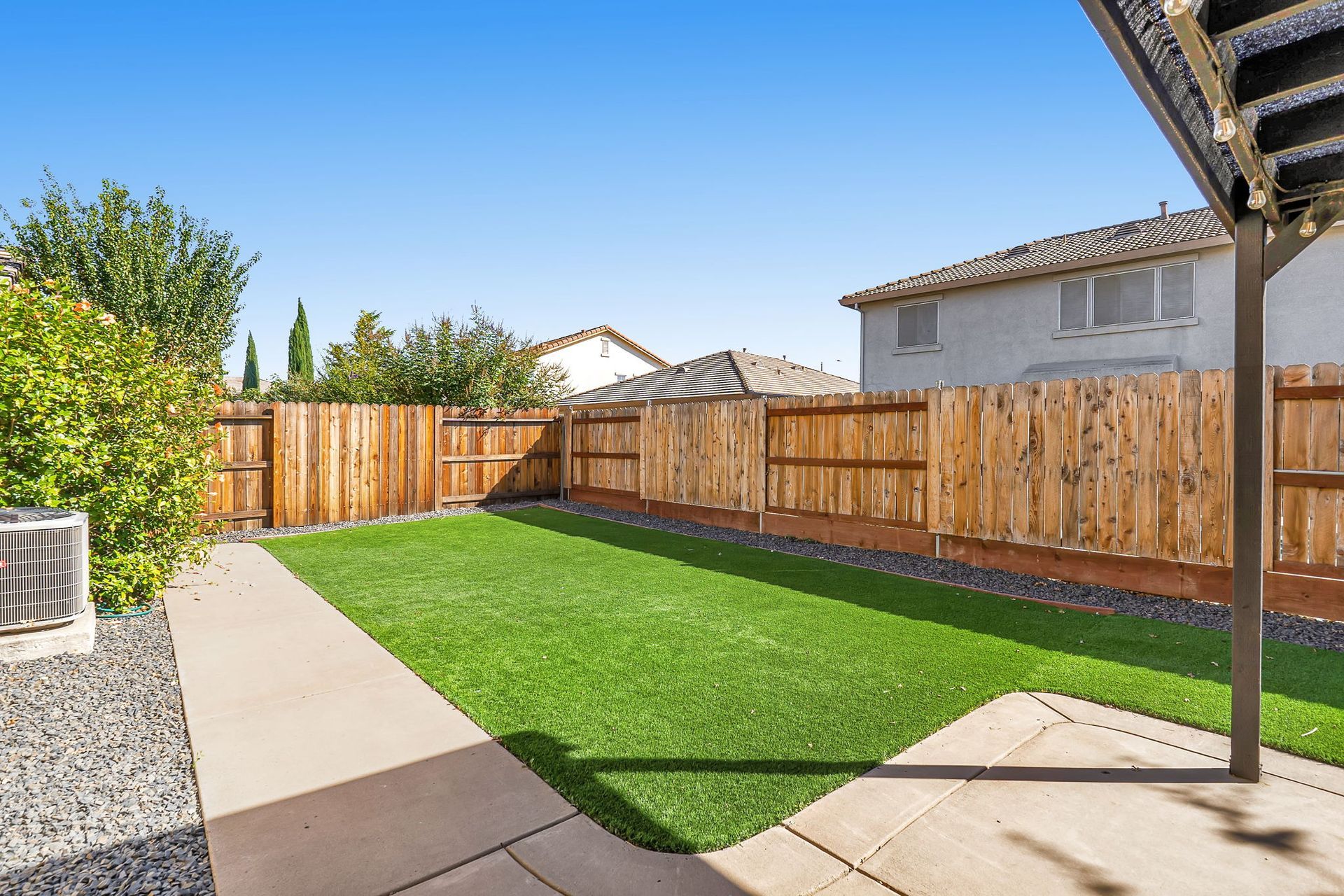 Backyard with green lawn, wooden fence, and concrete patio under a blue sky.