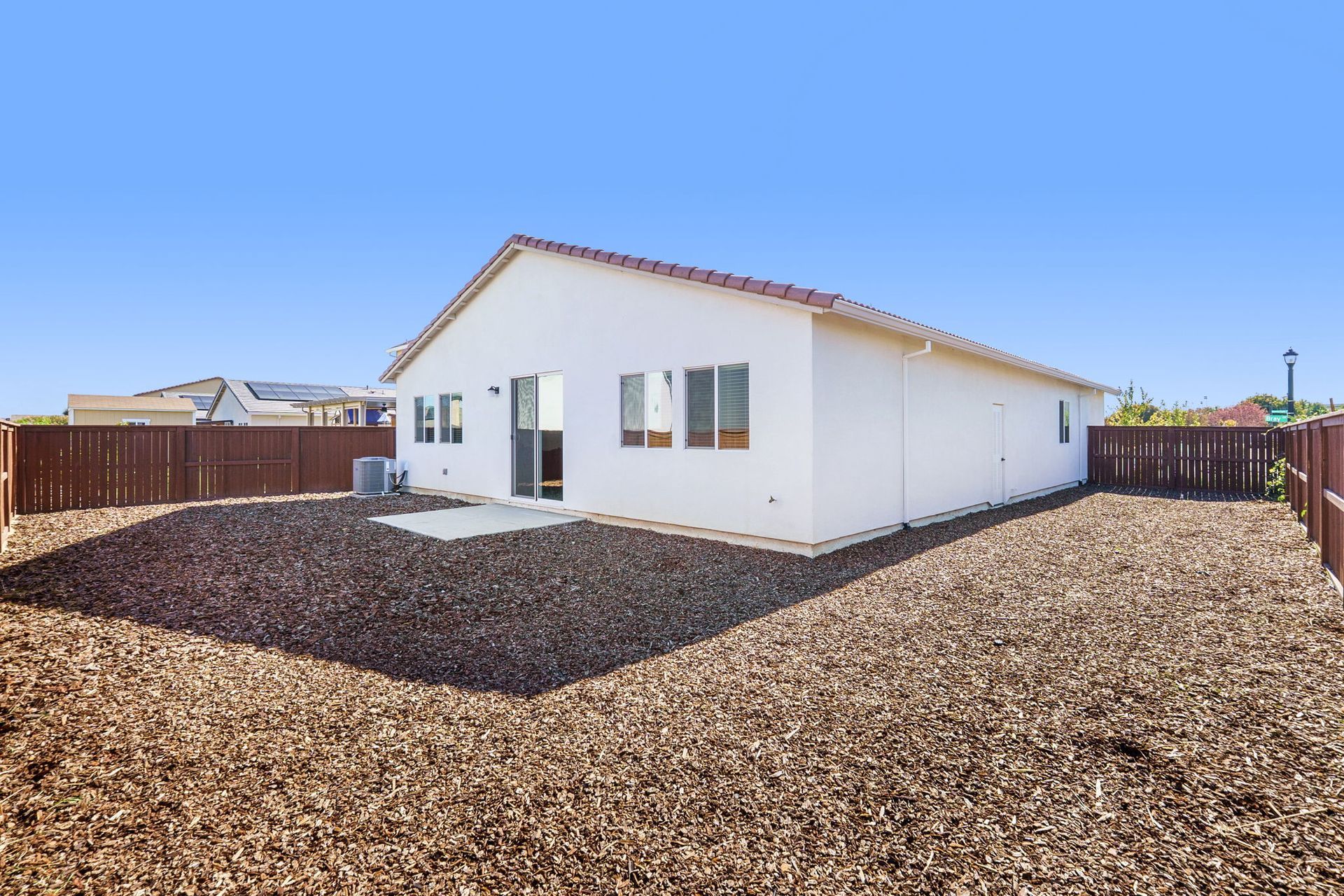 Backyard of a white house with brown mulch and a brown fence under a blue sky.