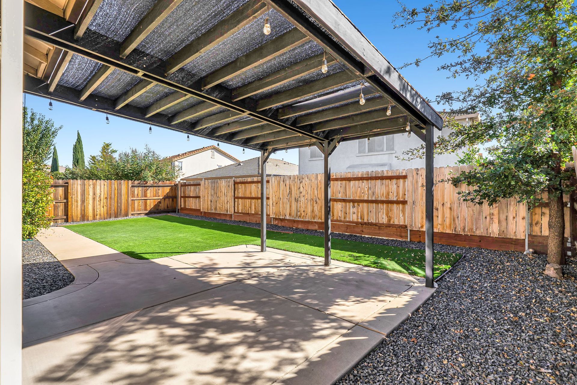 Backyard patio with a covered seating area, lawn, and wooden fence on a sunny day.