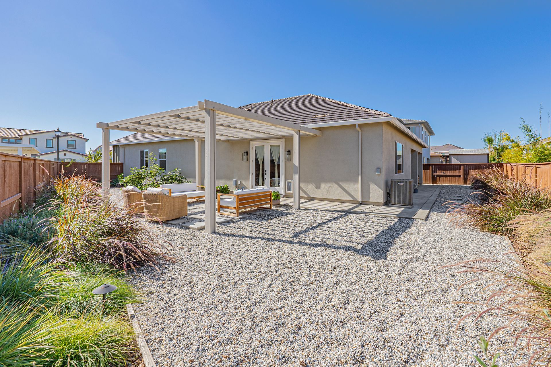 Backyard with gravel patio, pergola, seating, and stucco house under a blue sky.
