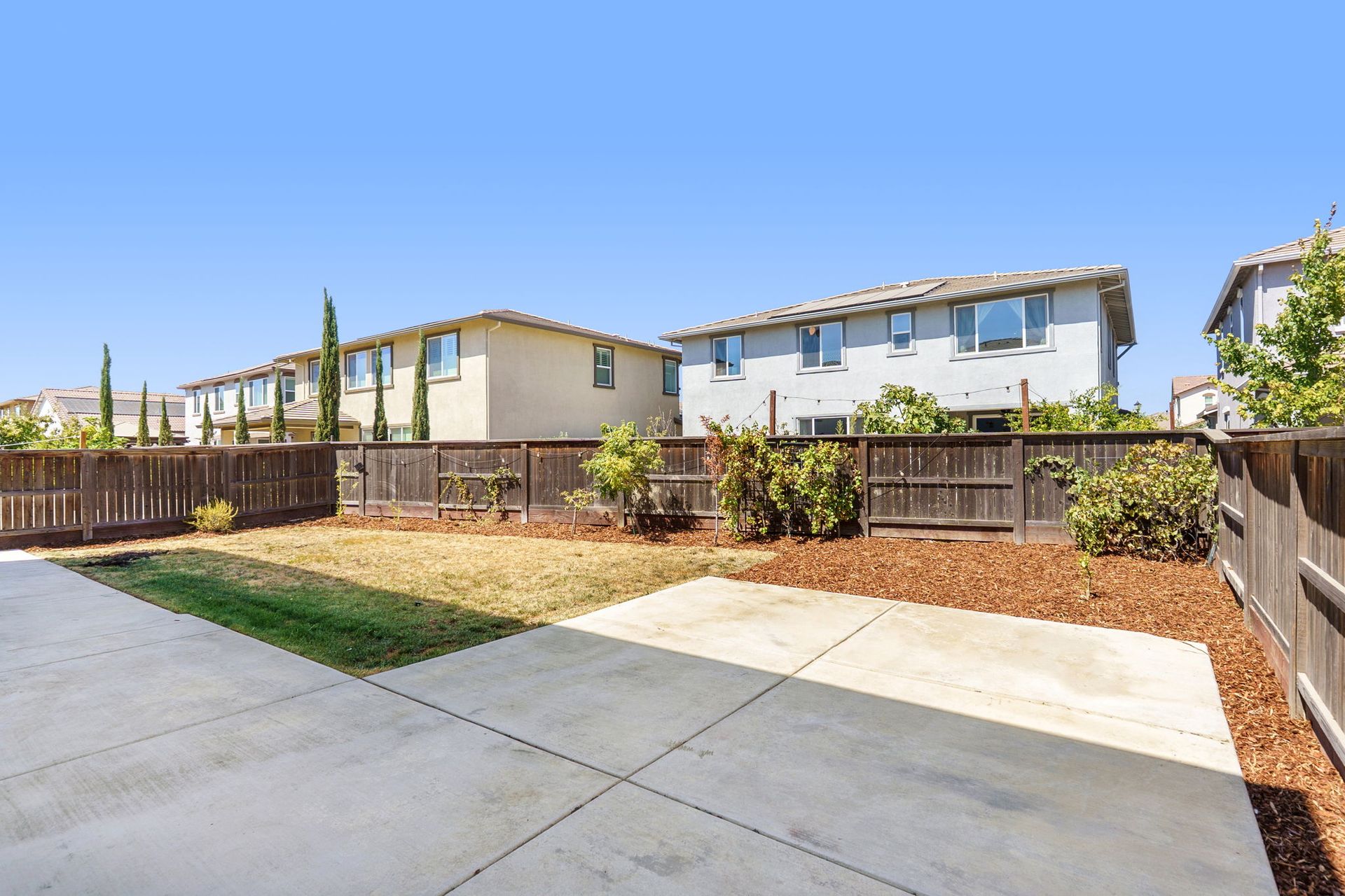 Backyard with concrete patio, grass, and wooden fence; houses in background under blue sky.
