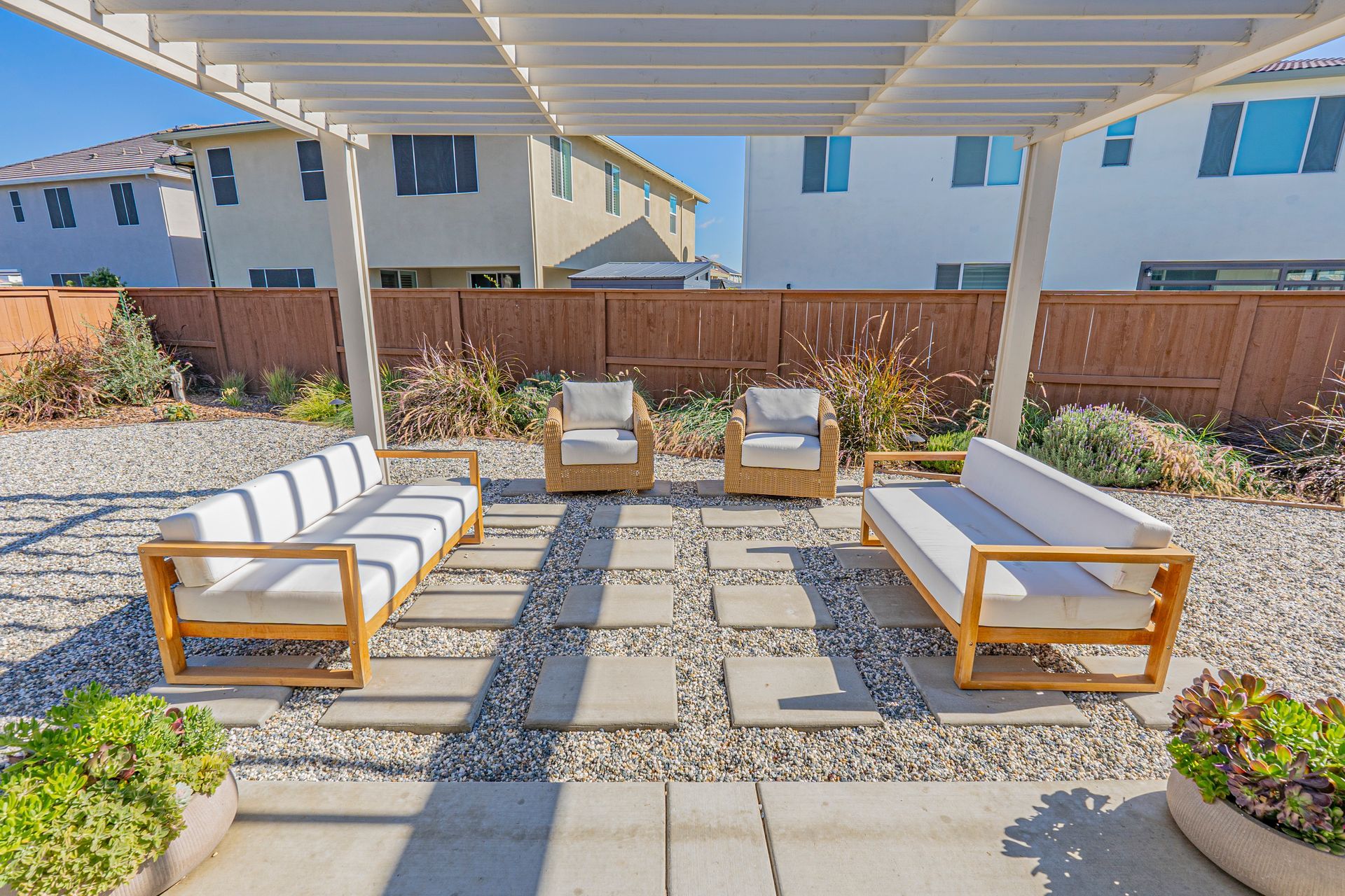 Backyard patio with wooden furniture under a pergola, gray stone pathway, and gravel.