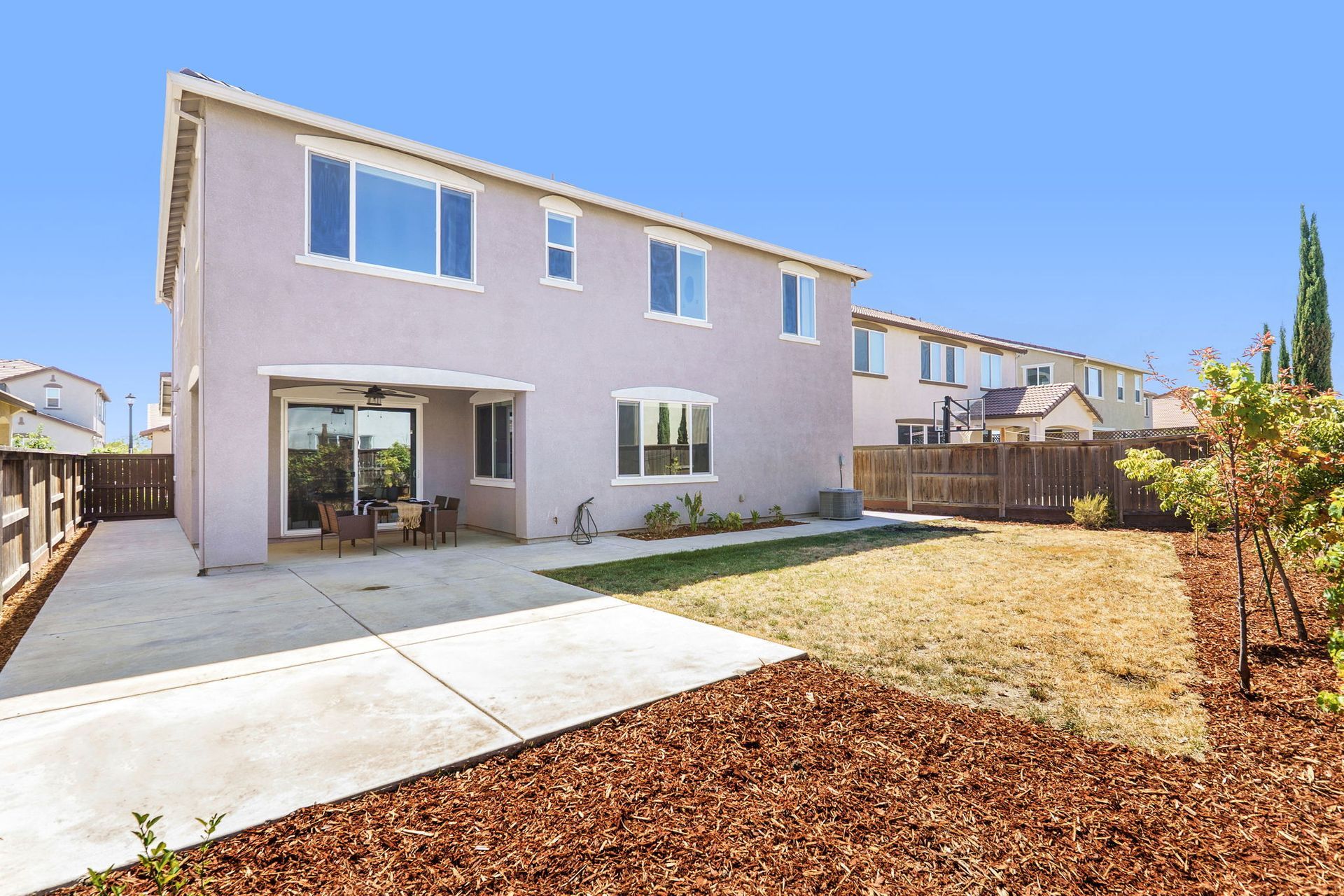 Backyard of a two-story house with patio, lawn, and wood-chip landscaping against a blue sky.