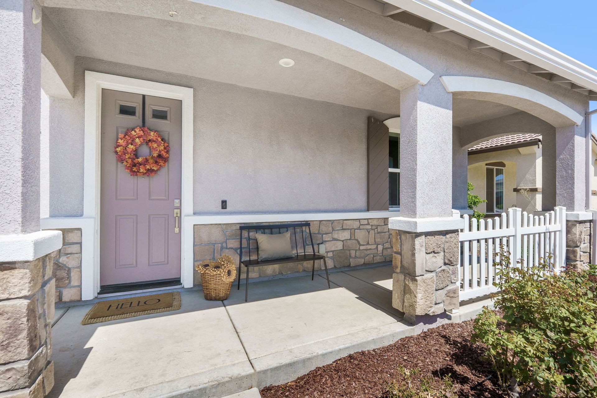 Front porch of a house with light grey exterior, columns with stone accents, pink door with wreath, bench, and picket fence.
