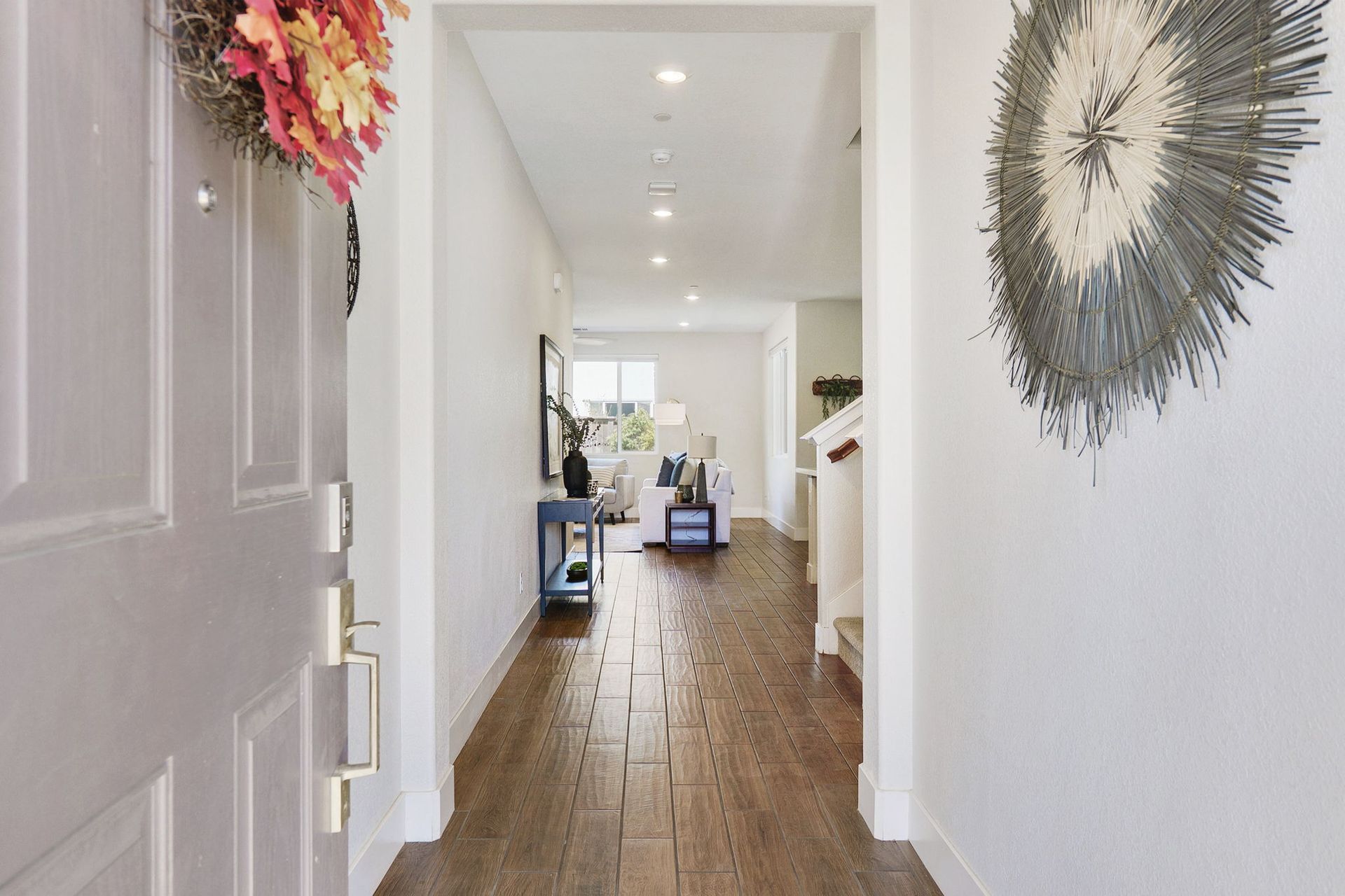 Open doorway into a bright home hallway with wood flooring. A decorative wreath hangs on the door.