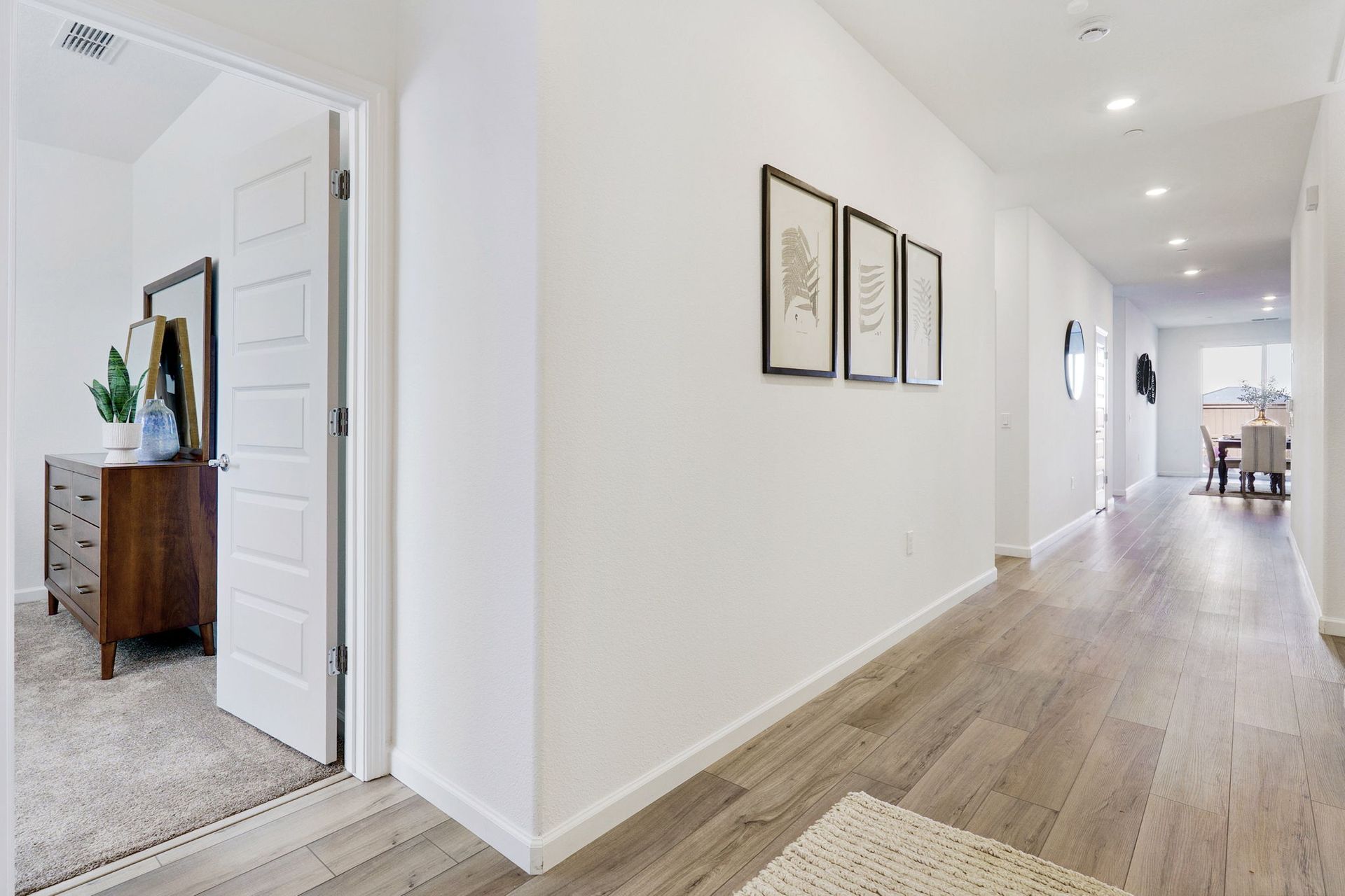 Hallway with wood floors, white walls, art, and an open doorway to a bedroom.
