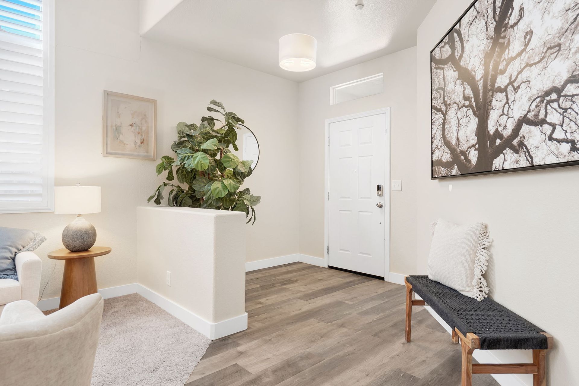Entryway with a bench, art, and a large plant. Light wood floors, white walls, and a pendant light.