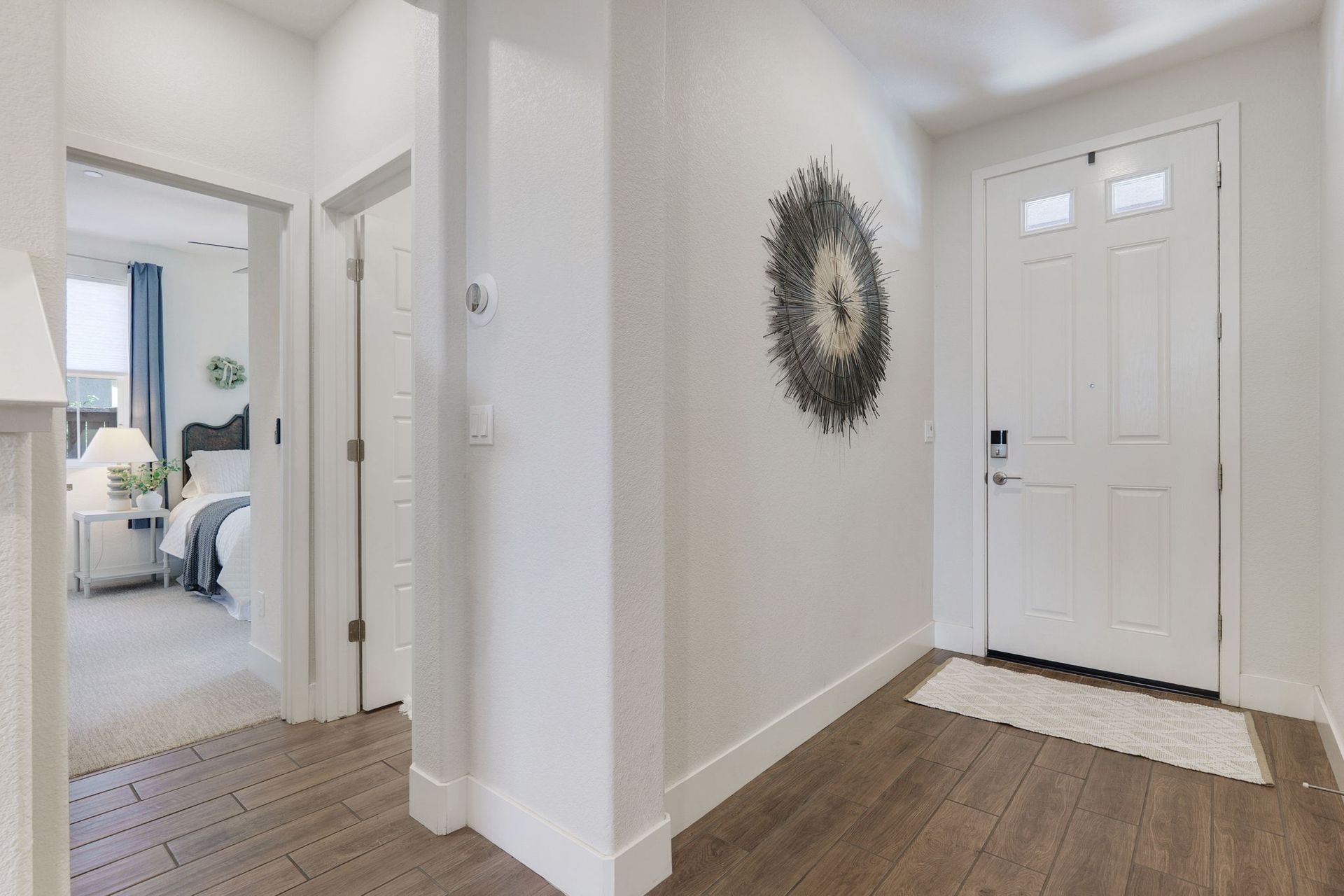 Hallway with white walls, wood floors, and a front door. Artwork and a bedroom are visible.
