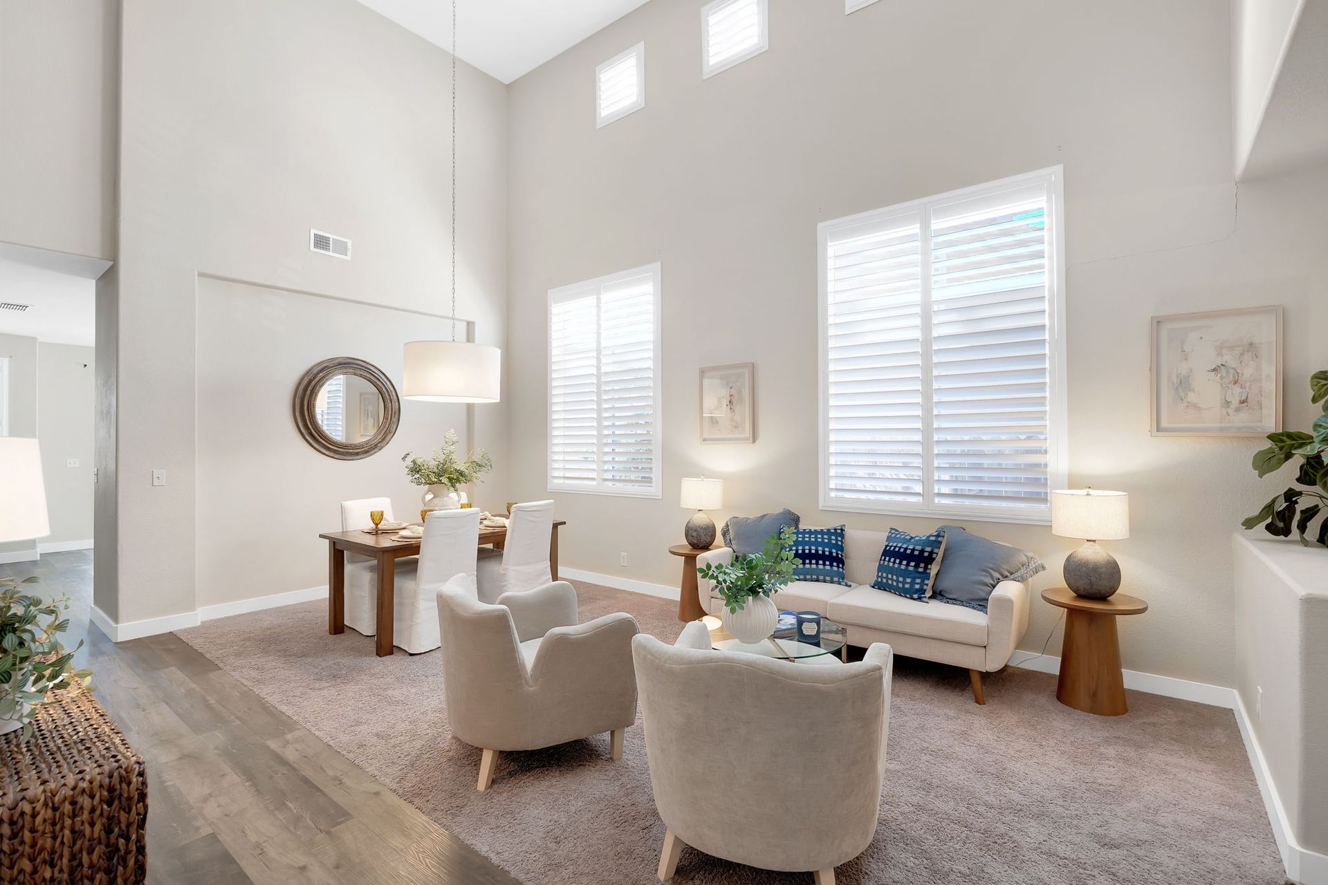 Living room with high ceilings, beige walls, windows, and light-colored furniture on a brown carpet.