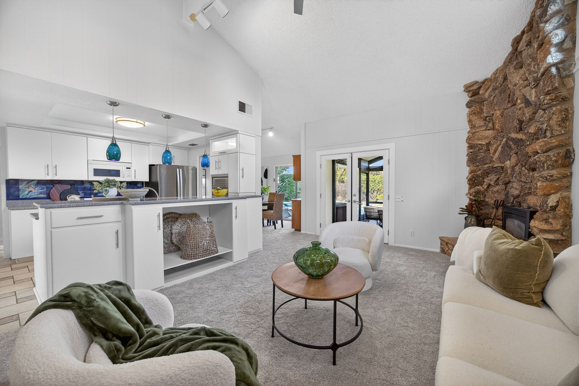 Cozy living room with open kitchen, white walls, and stone fireplace. Two armchairs and a sofa are present.