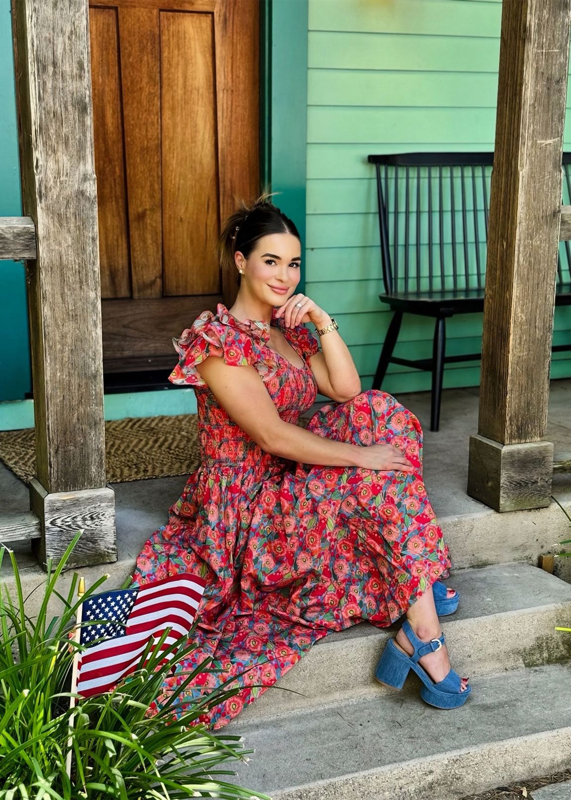 Woman in floral dress, blue shoes, sitting on steps with American flag. Green house backdrop.