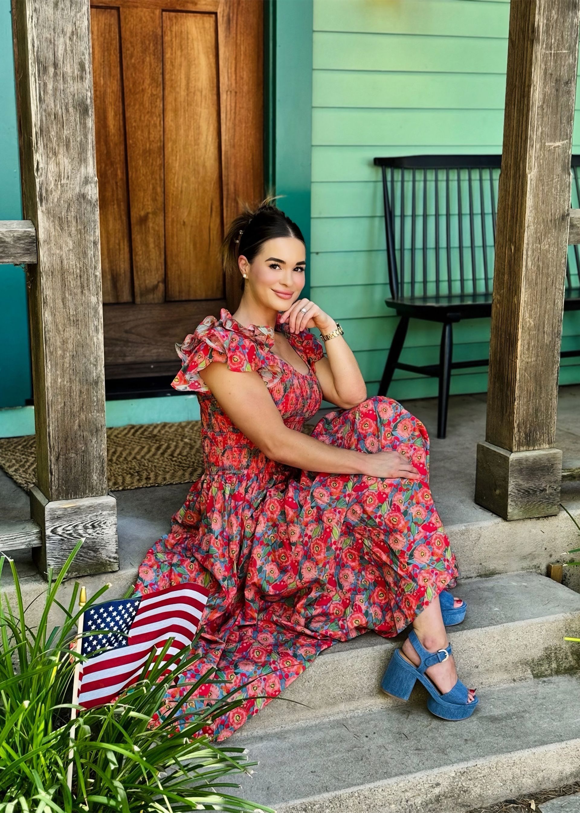 Woman in floral dress, blue shoes, sitting on steps with American flag. Green house backdrop.