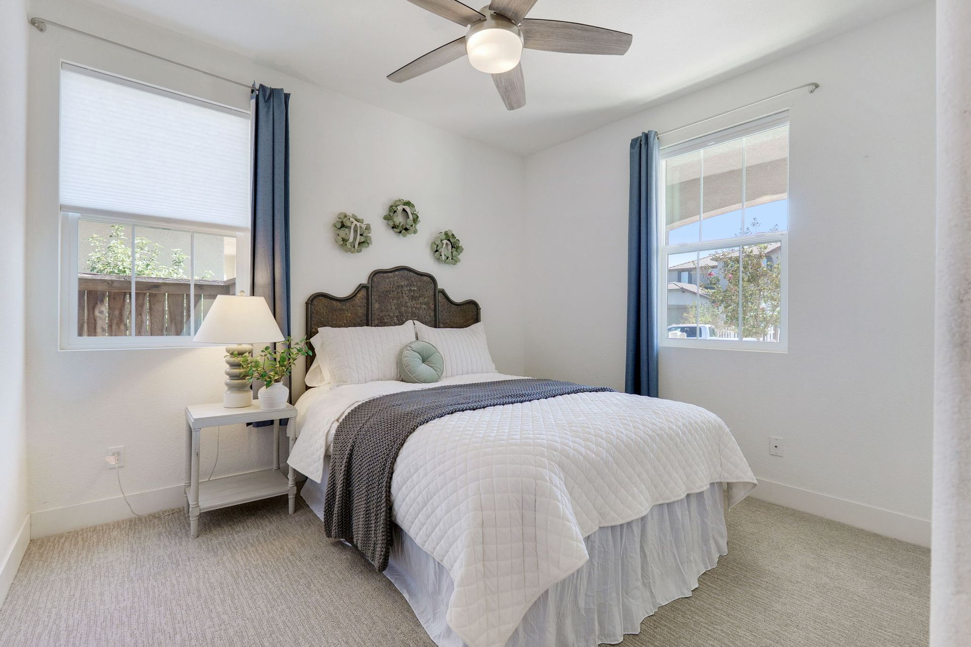 Bedroom with bed, nightstand, windows, and ceiling fan. White walls, carpet, and bedding. Blue curtains.