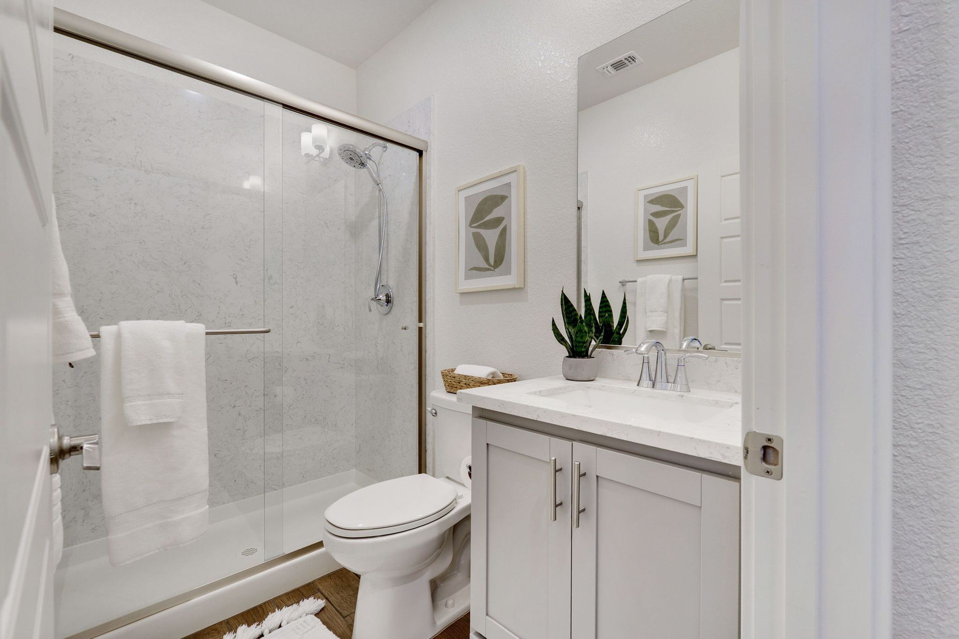 White bathroom with a shower, toilet, and vanity. Decorative wall art and a potted plant are on the counter.