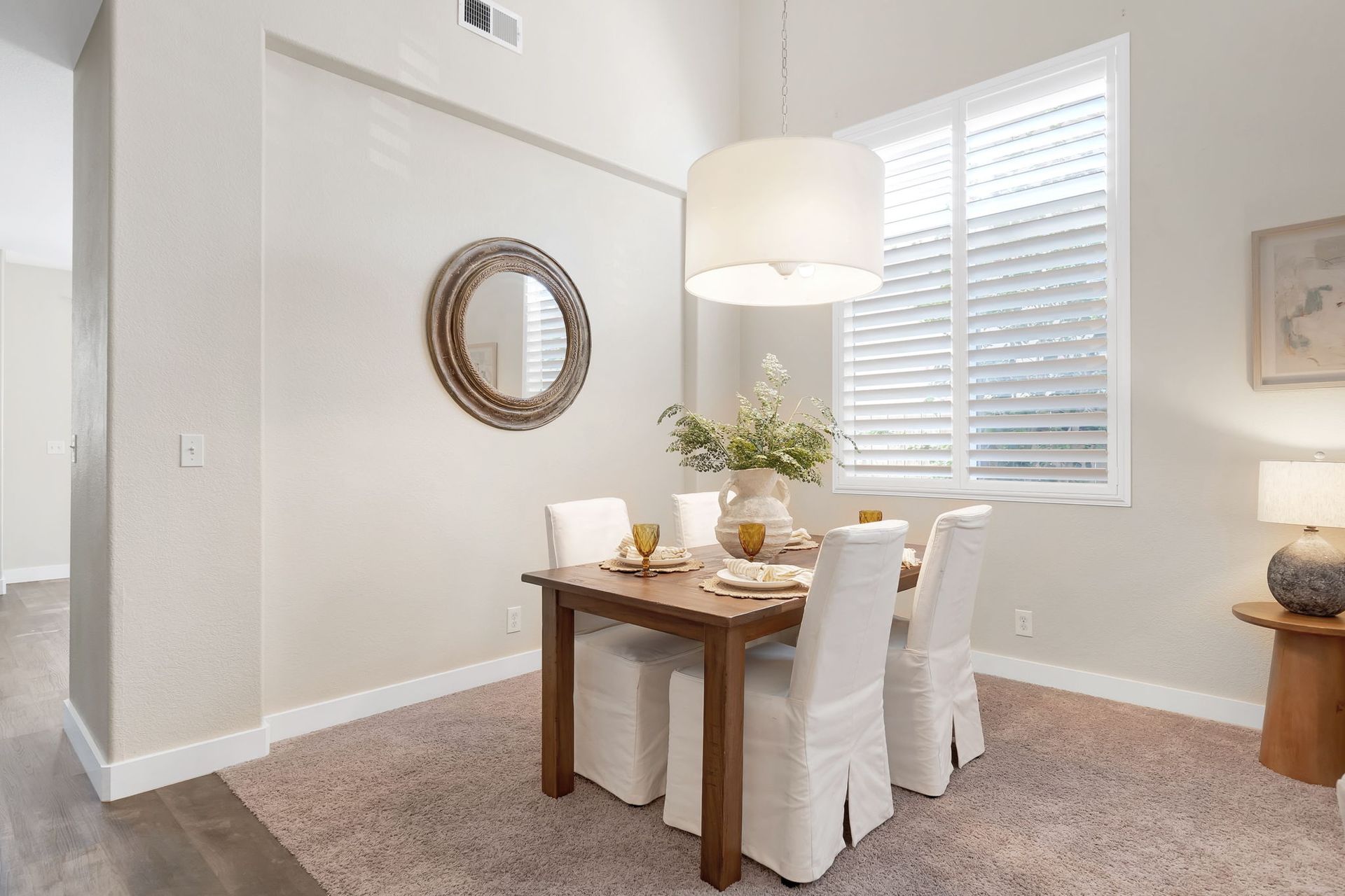 Dining area: wooden table, white chairs, round mirror, large light, window with shutters, and floral arrangement.