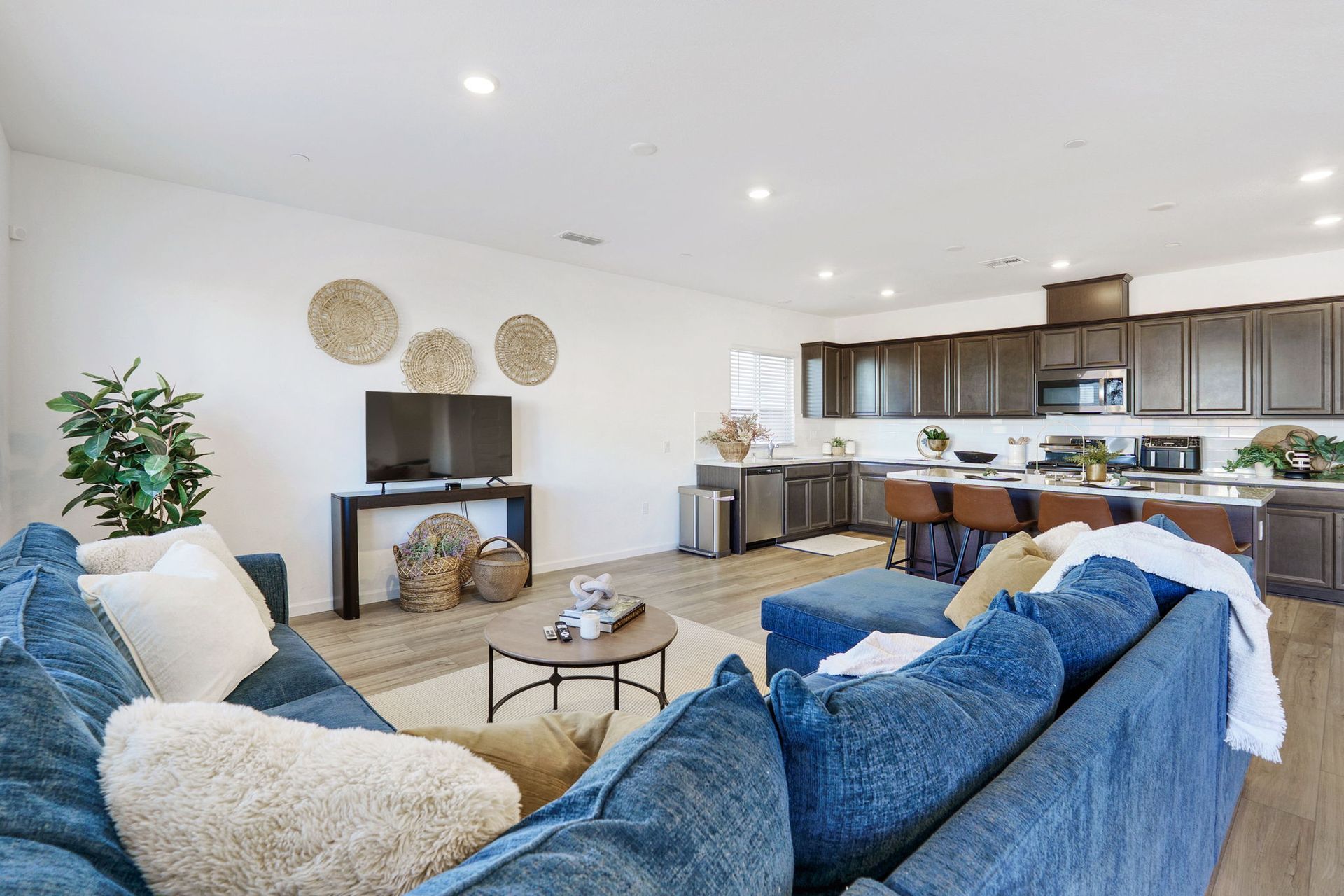 Open-concept living space with blue sofa, kitchen with dark cabinets, and woven wall decor.