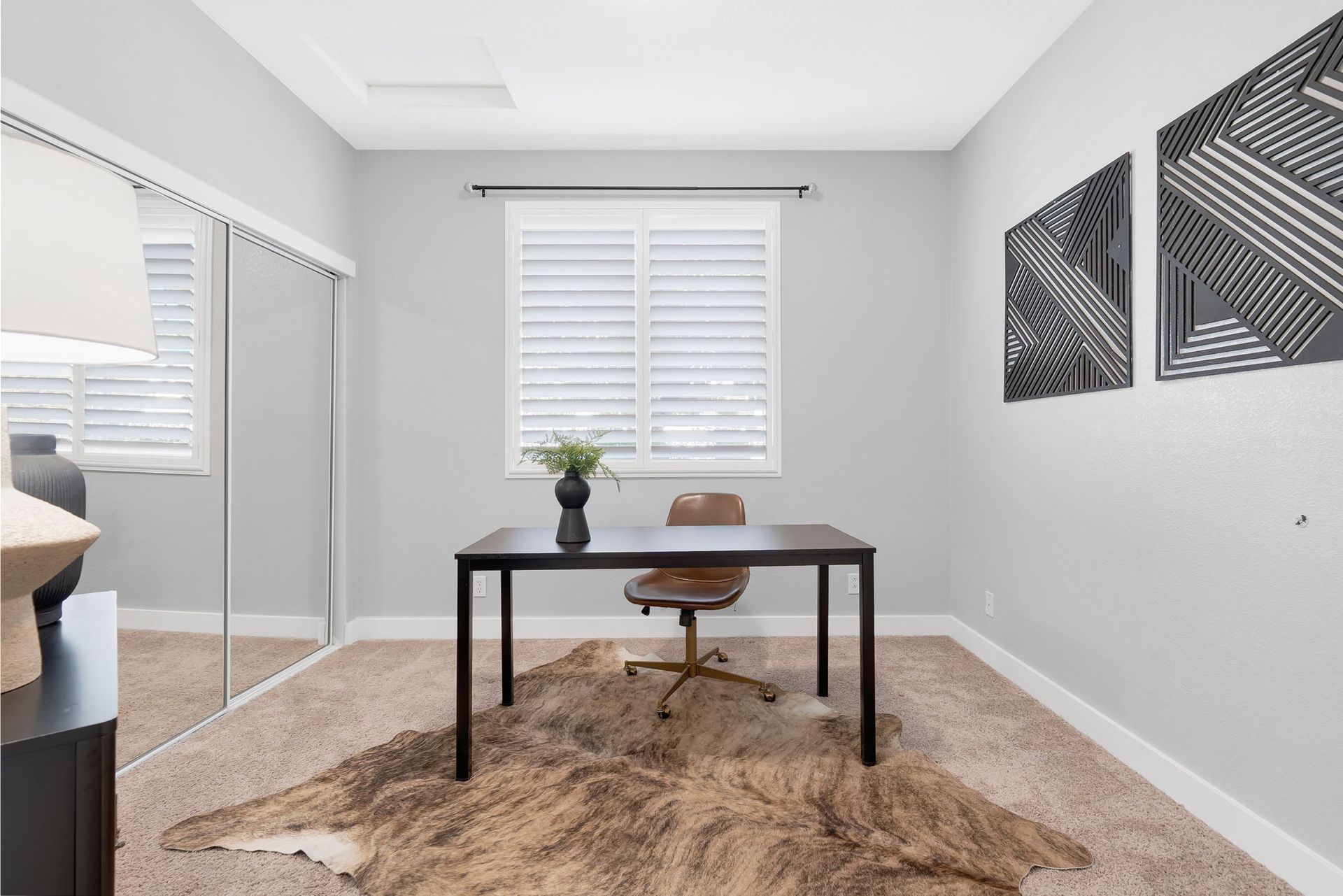 Office space with a black desk, brown chair, and cowhide rug, with gray walls, window with shutters, and art.