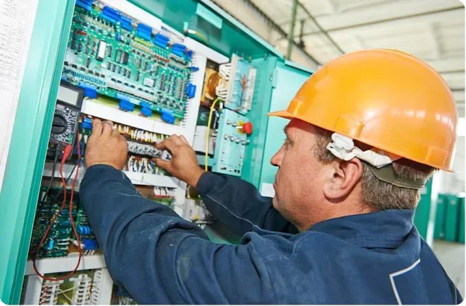 Technician in a hard hat inspects wiring and circuit boards inside an open industrial control panel.