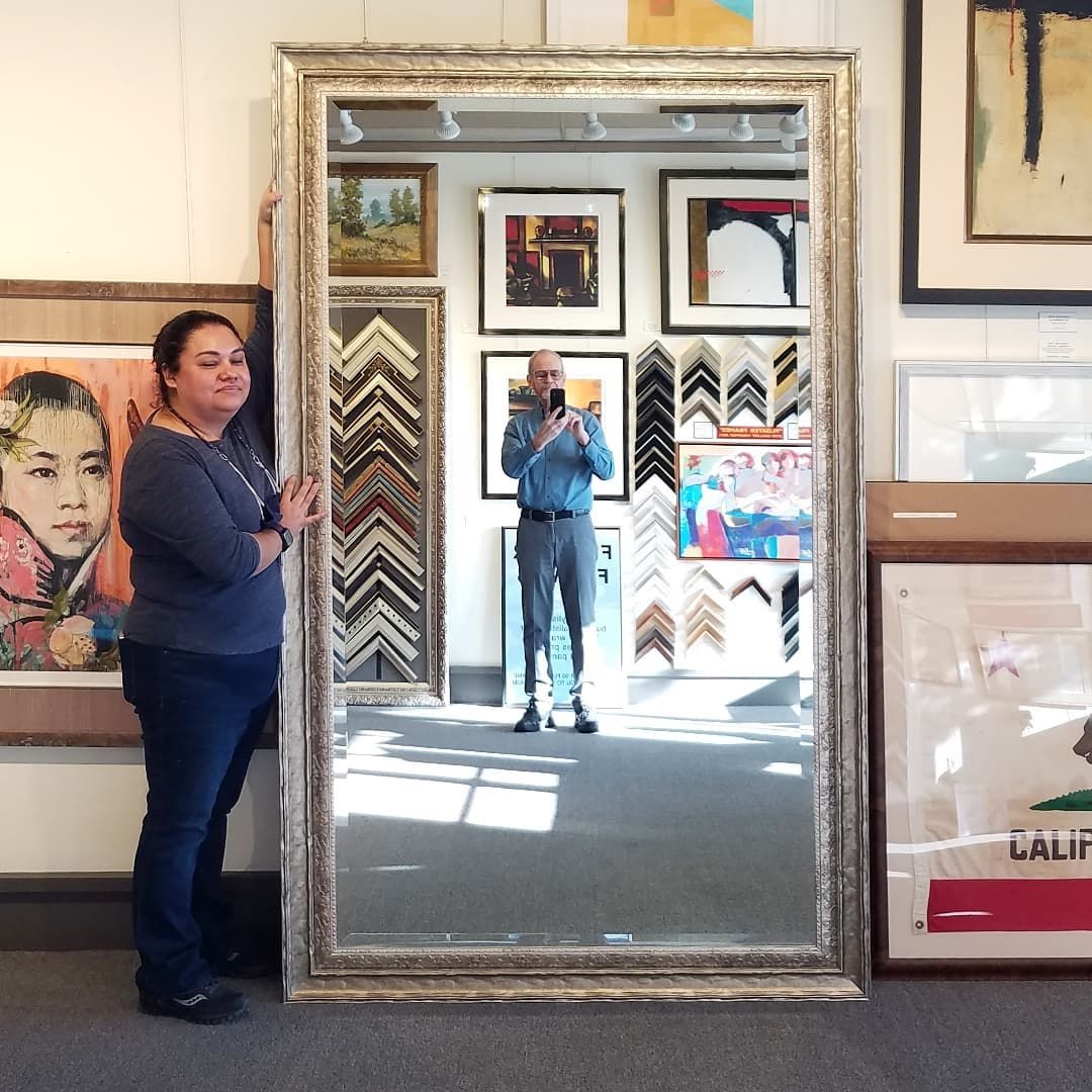 Woman stands beside a tall ornate mirror in an art shop, with her reflection and camera visible.