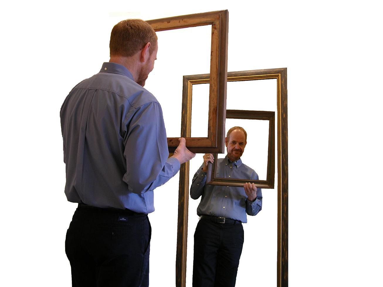 Man holding a large wooden frame, seen reflected in a mirror-like frame within a white room