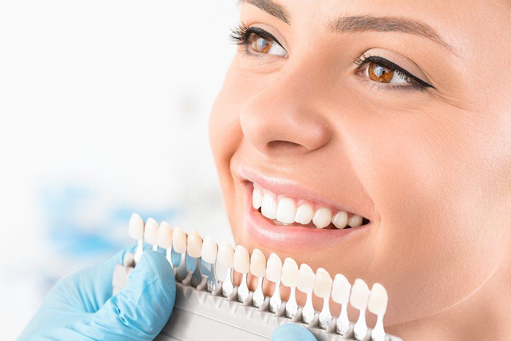 Woman smiling, holding teeth shade guide during dental checkup.