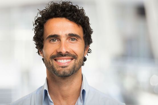 Man with curly dark hair and a beard, smiling. Wearing a light blue shirt.