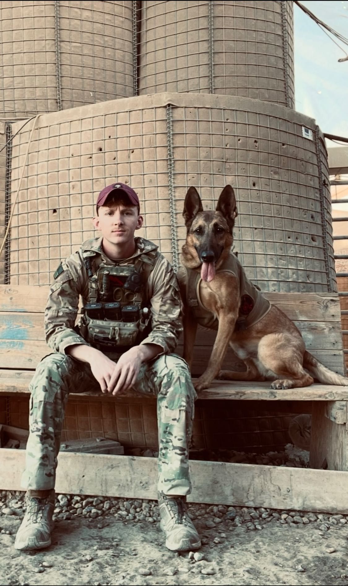 A soldier in uniform sits on a wooden bench next to a German Shepherd dog, with large cylindrical structures behind them.