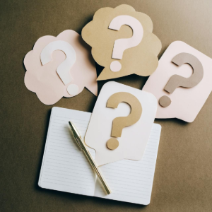 A gold pen rests on an open notebook surrounded by four colorful speech bubbles, each featuring a large question mark.