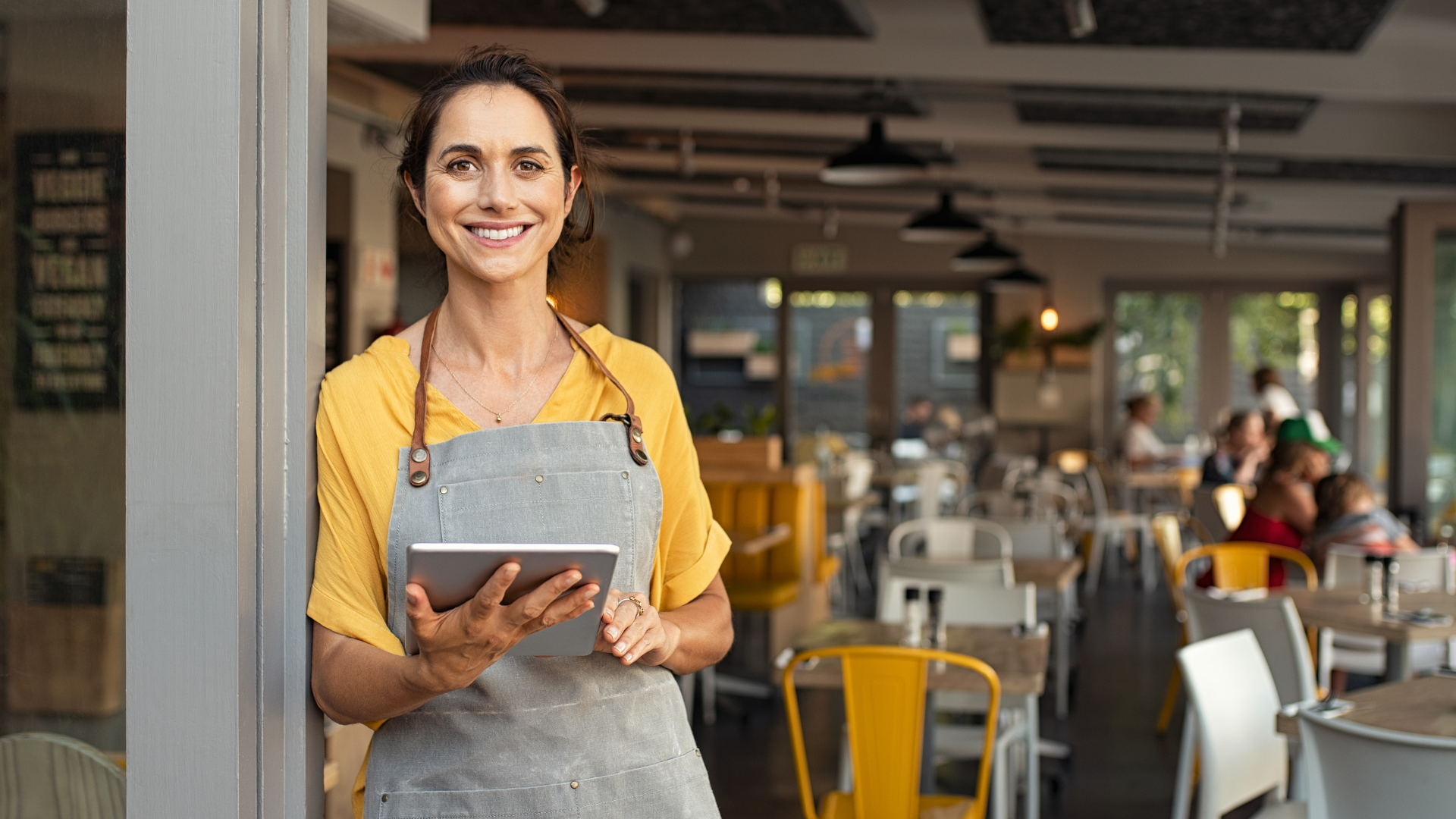 A woman is standing in the doorway of a restaurant holding a tablet.