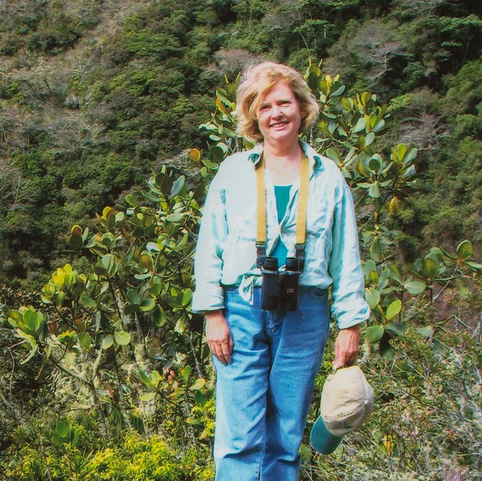 a woman wearing binoculars and a hat stands in the woods