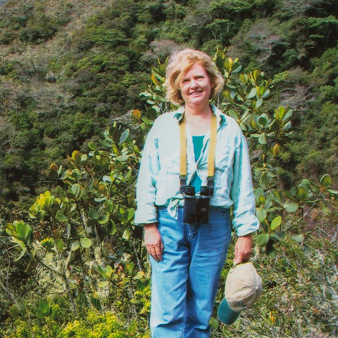 a woman wearing binoculars and a hat stands in the woods
