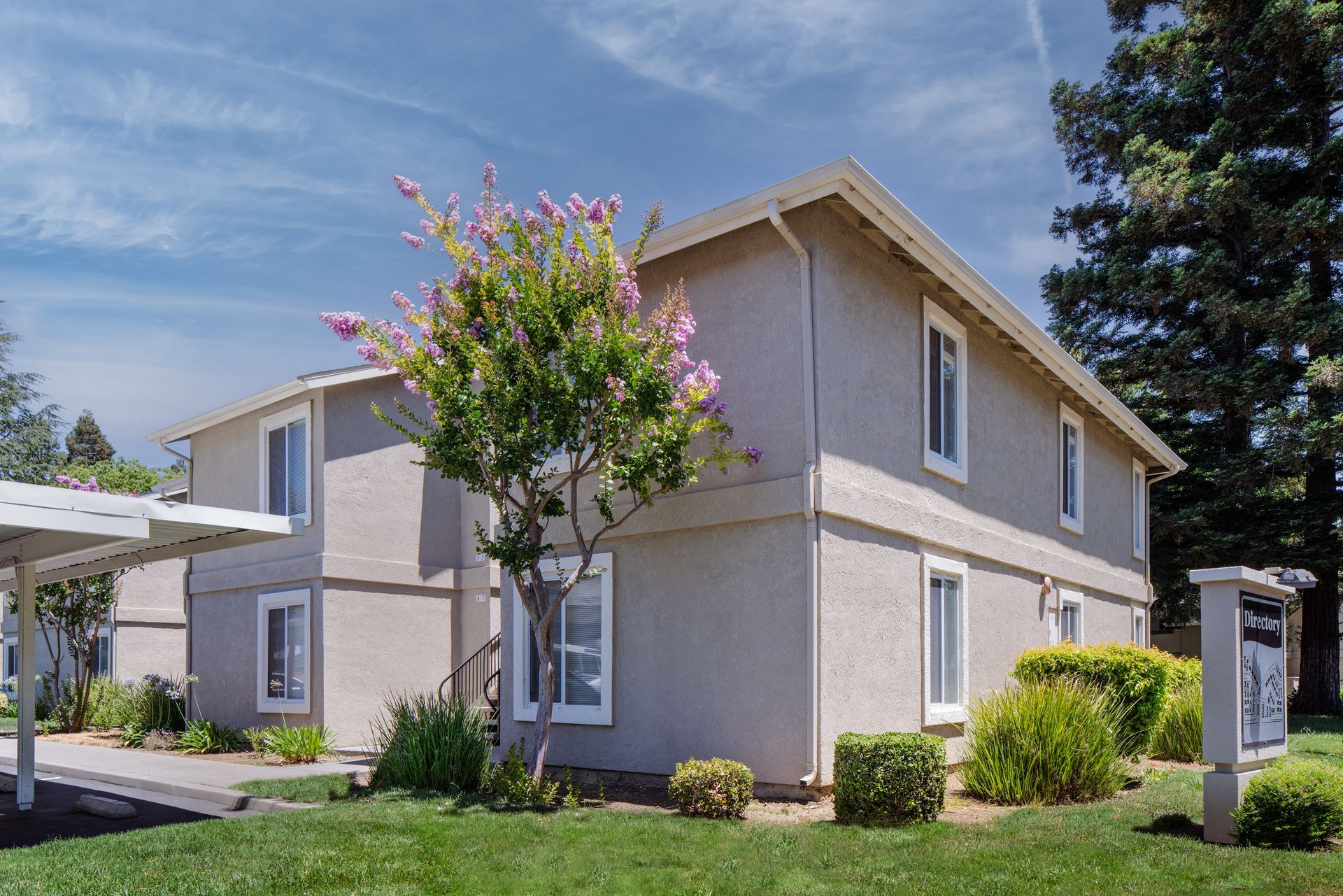 Two-story apartment building with beige exterior, white-framed windows, and green lawn under a blue sky.