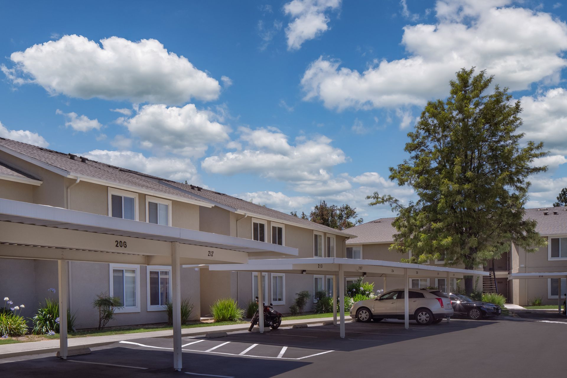 Cars parked under a carport next to a two-story apartment building on a sunny day.
