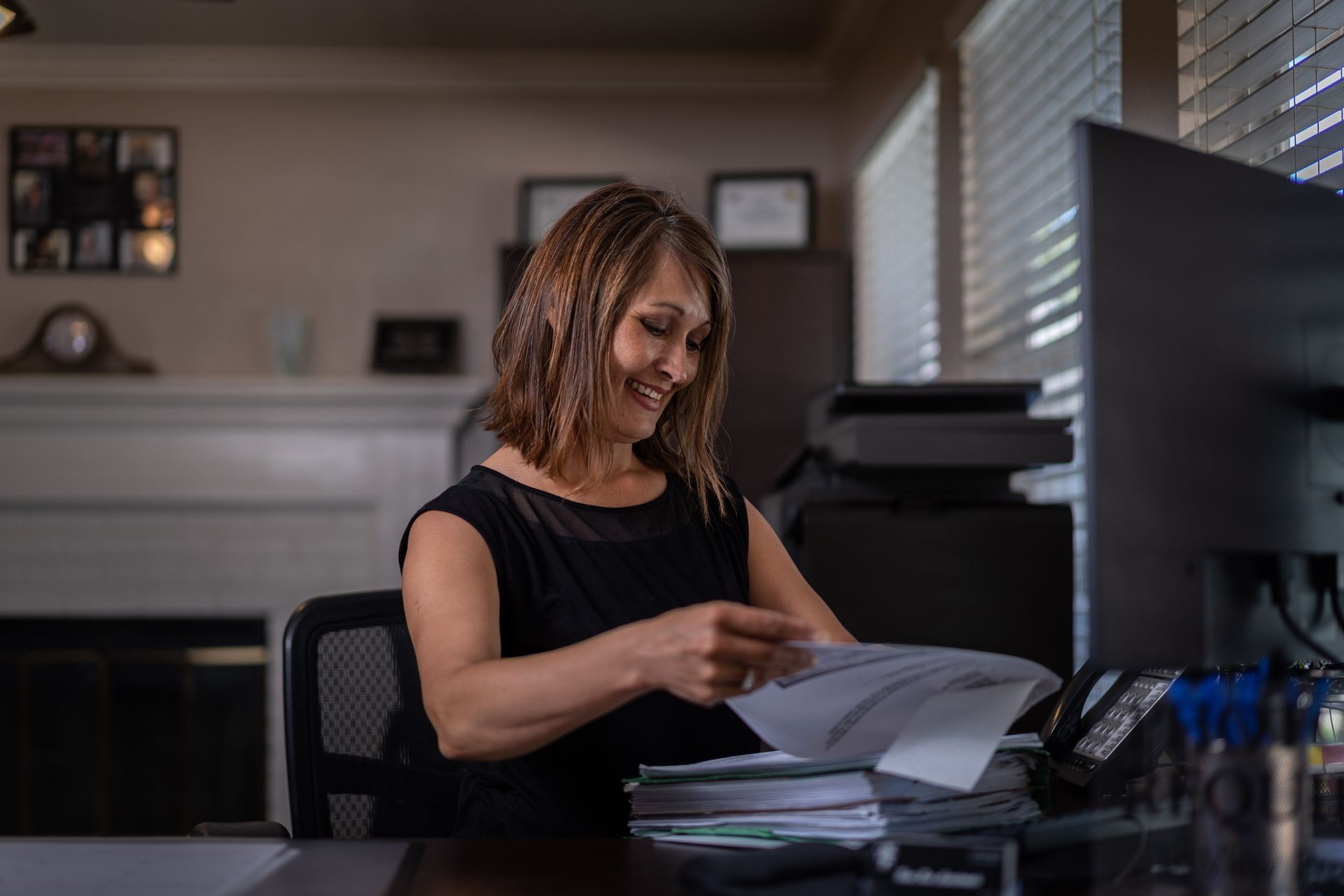 Woman in black top smiles while sorting papers at her desk.