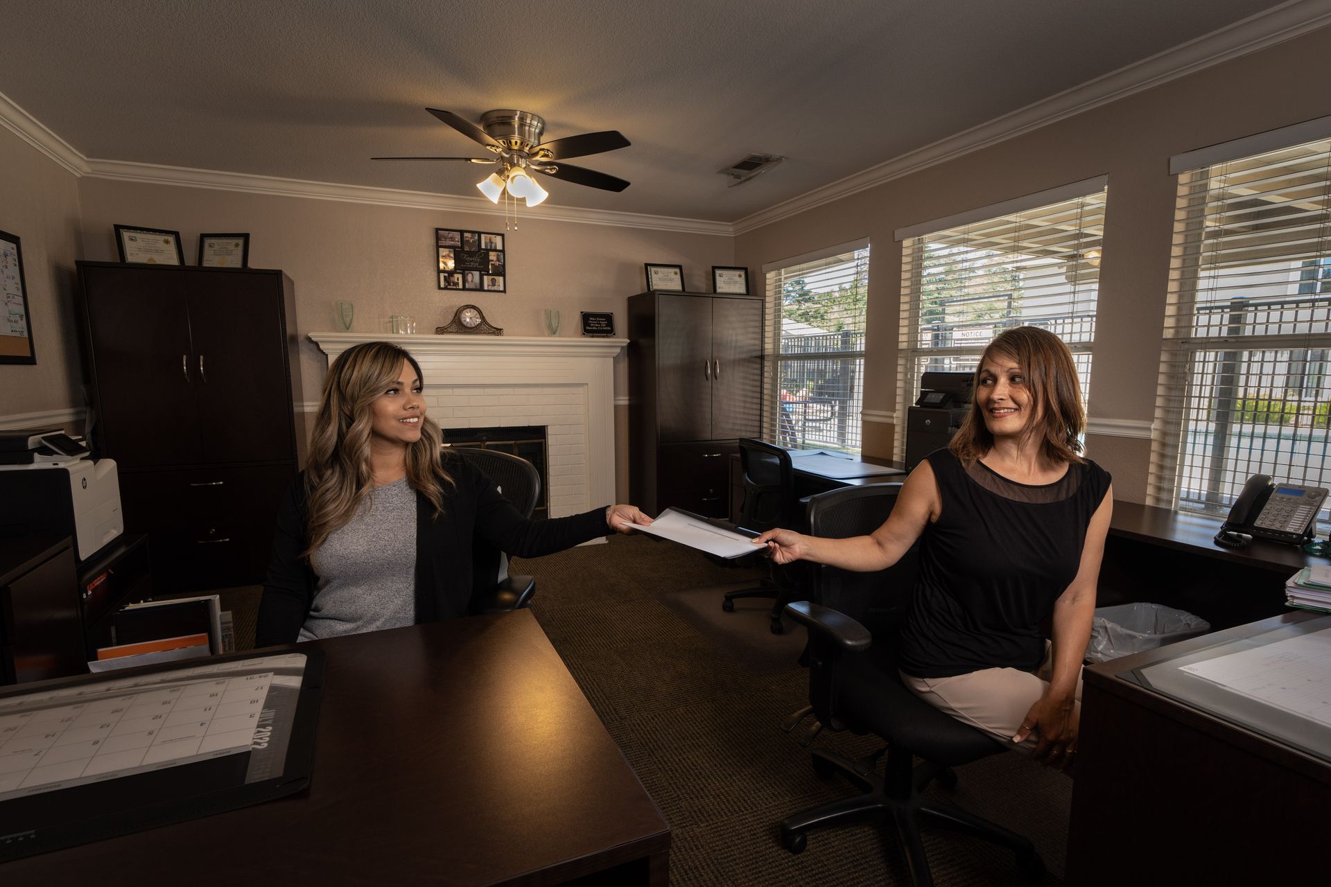 Two women in an office, one passing a document to the other, smiling.