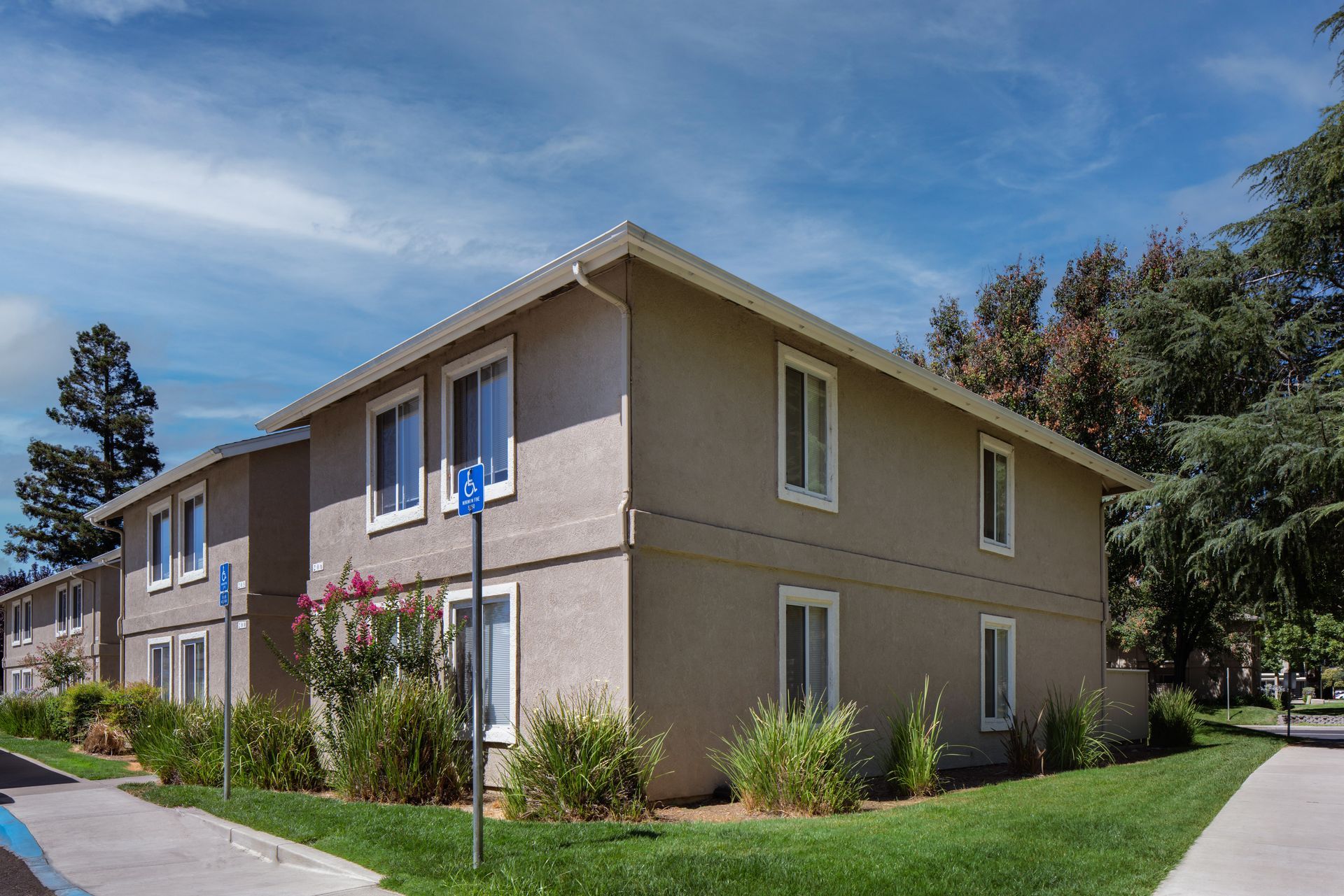 Tan two-story apartment building with white-framed windows, accessible parking sign, blue sky, and green lawn.