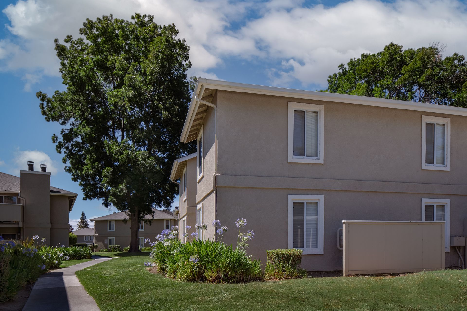 Two-story beige apartment building with white-framed windows; lawn and trees in a sunny setting.