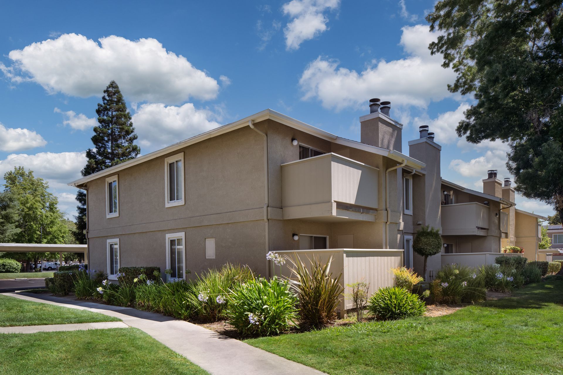 Two-story beige apartment building with balconies under a blue sky with clouds. Green grass and shrubs in the foreground.