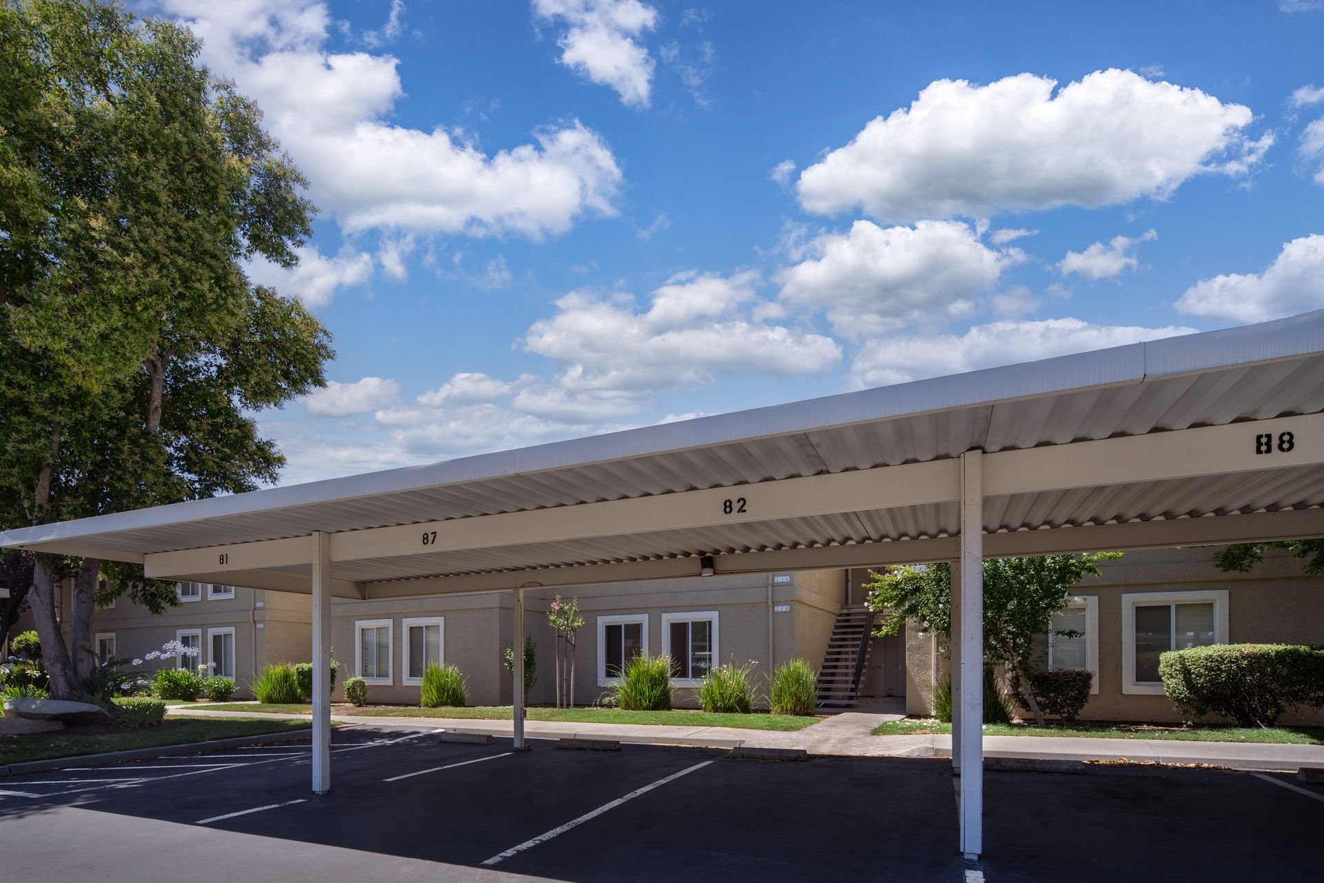 Carport sheltering parked cars in front of beige apartment buildings under a cloudy blue sky.