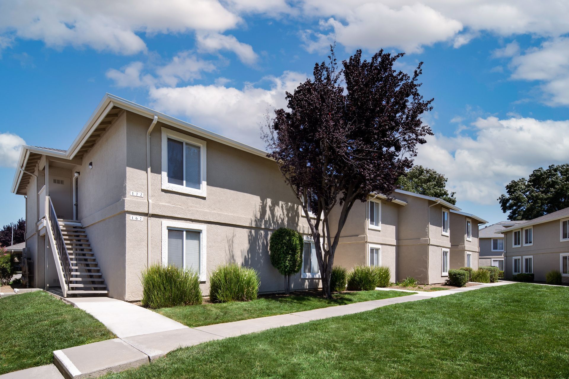 Apartment complex with green lawn, trees, and blue sky with clouds.